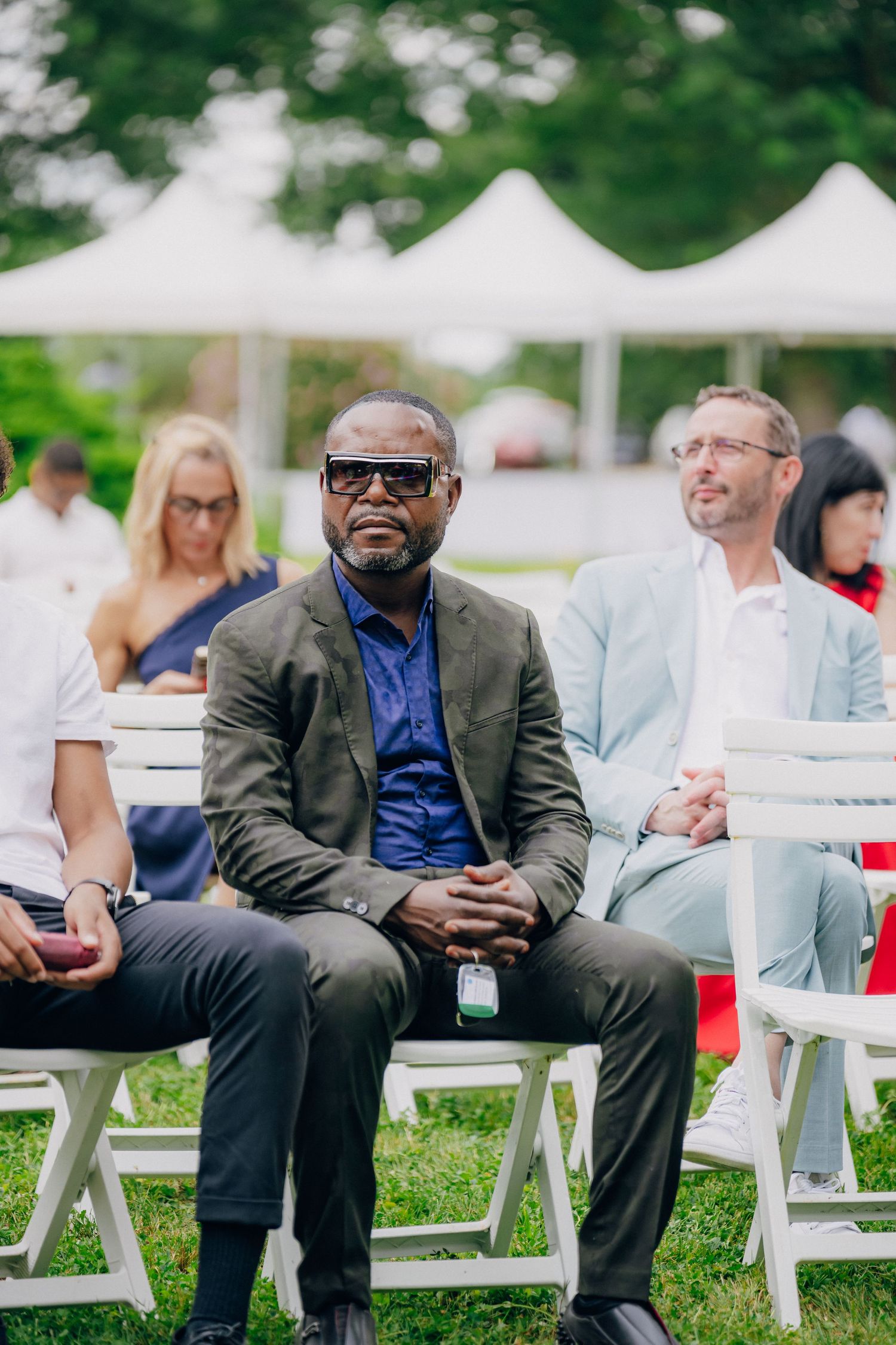 Guests seated in white folding chairs at an outdoor event with tents visible in the background.