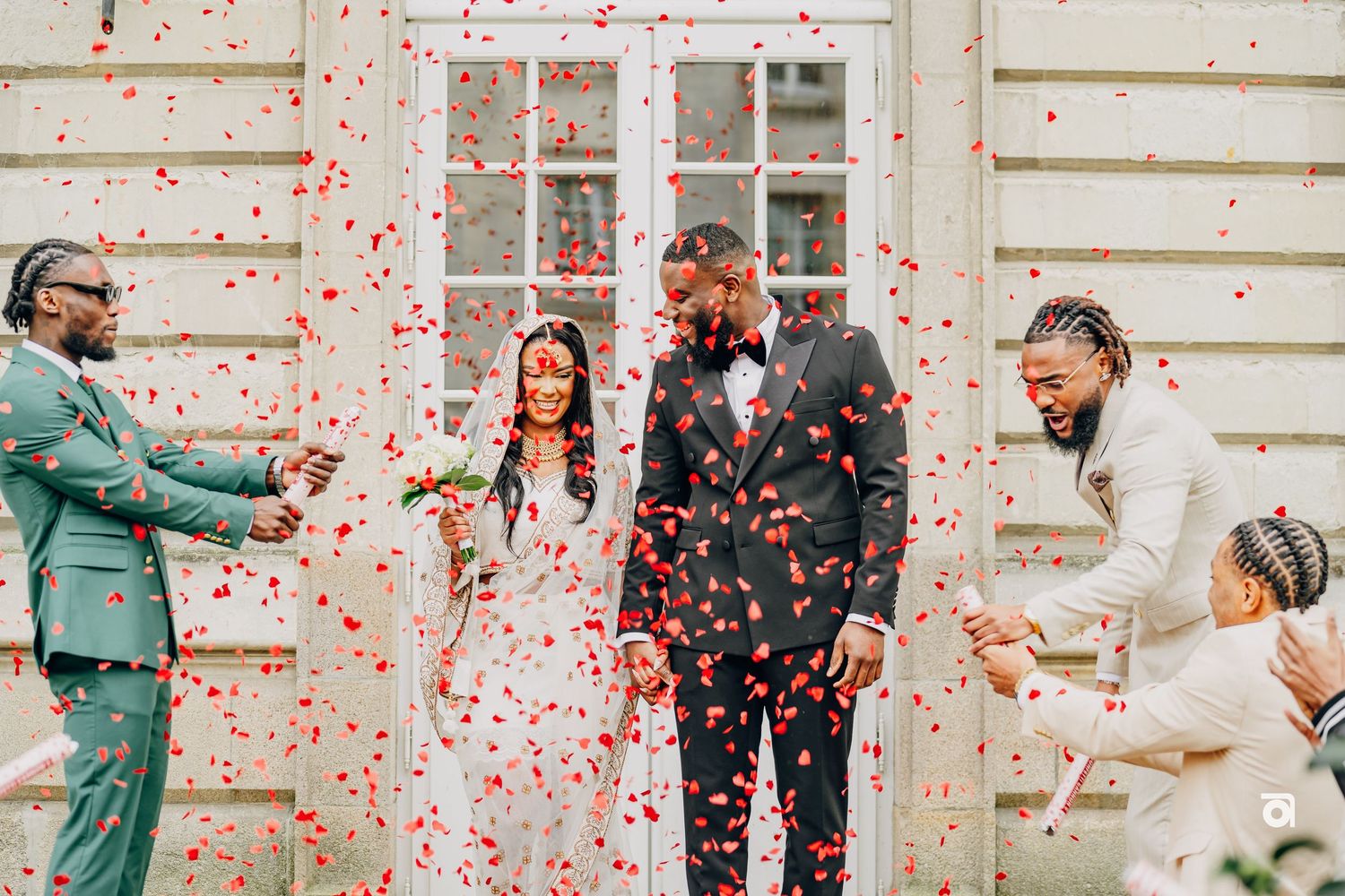 Wedding celebration outside with red confetti shower against white building backdrop.