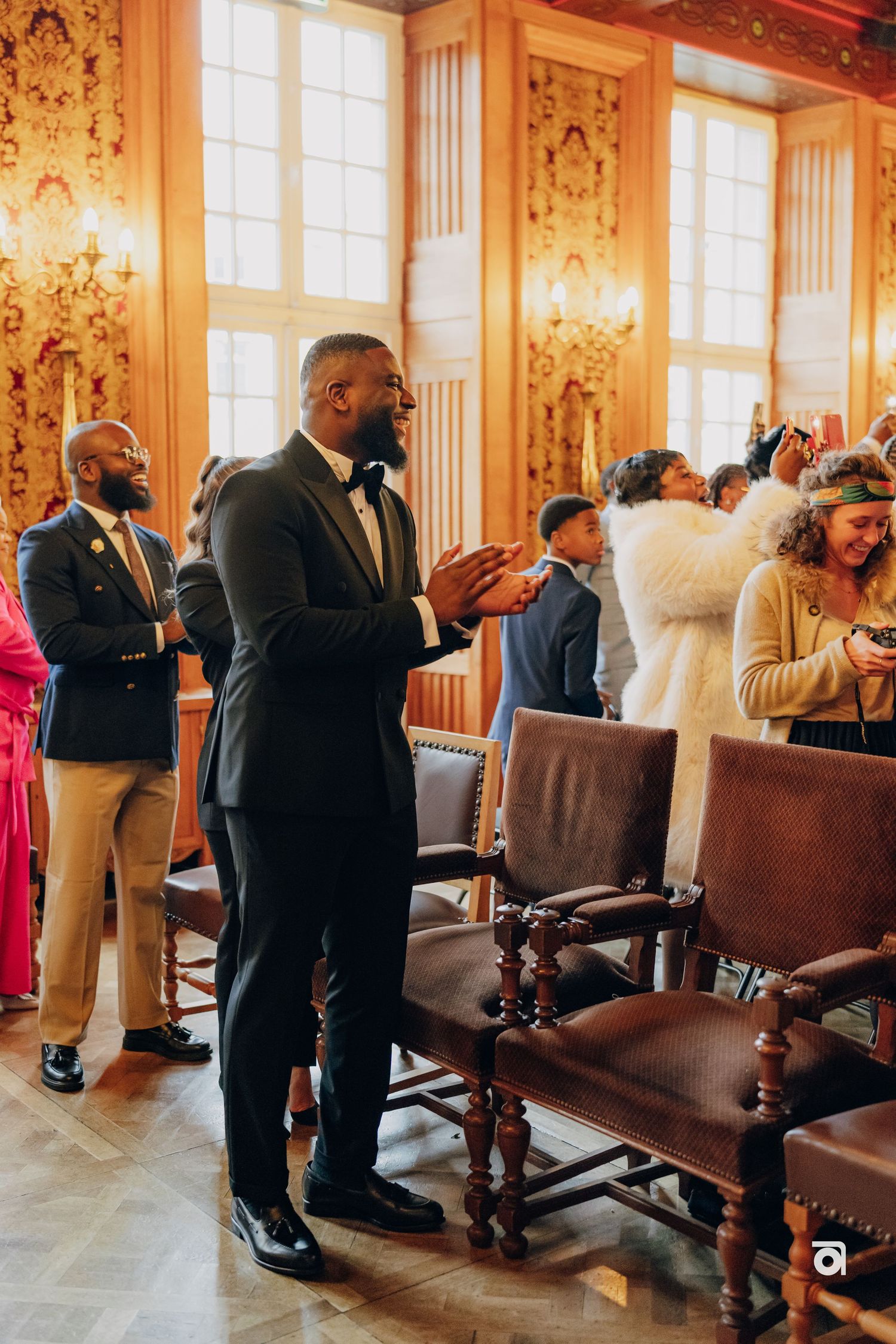 Wedding guests standing in an ornate ballroom with golden walls and large windows during a celebration.