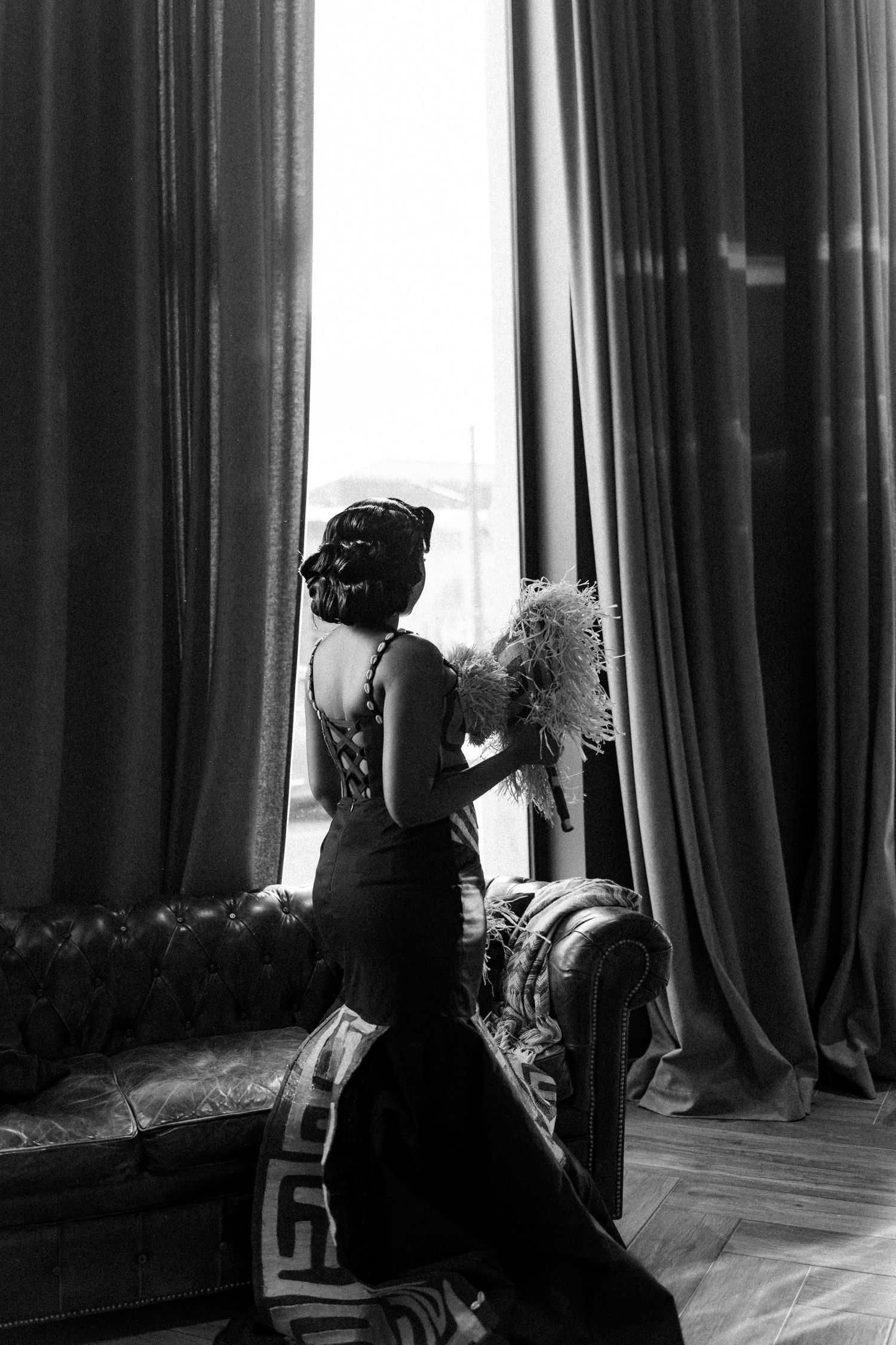 A vintage-style black and white photo of an elegant woman in a formal dress gazing out a tall window with flowing curtains.