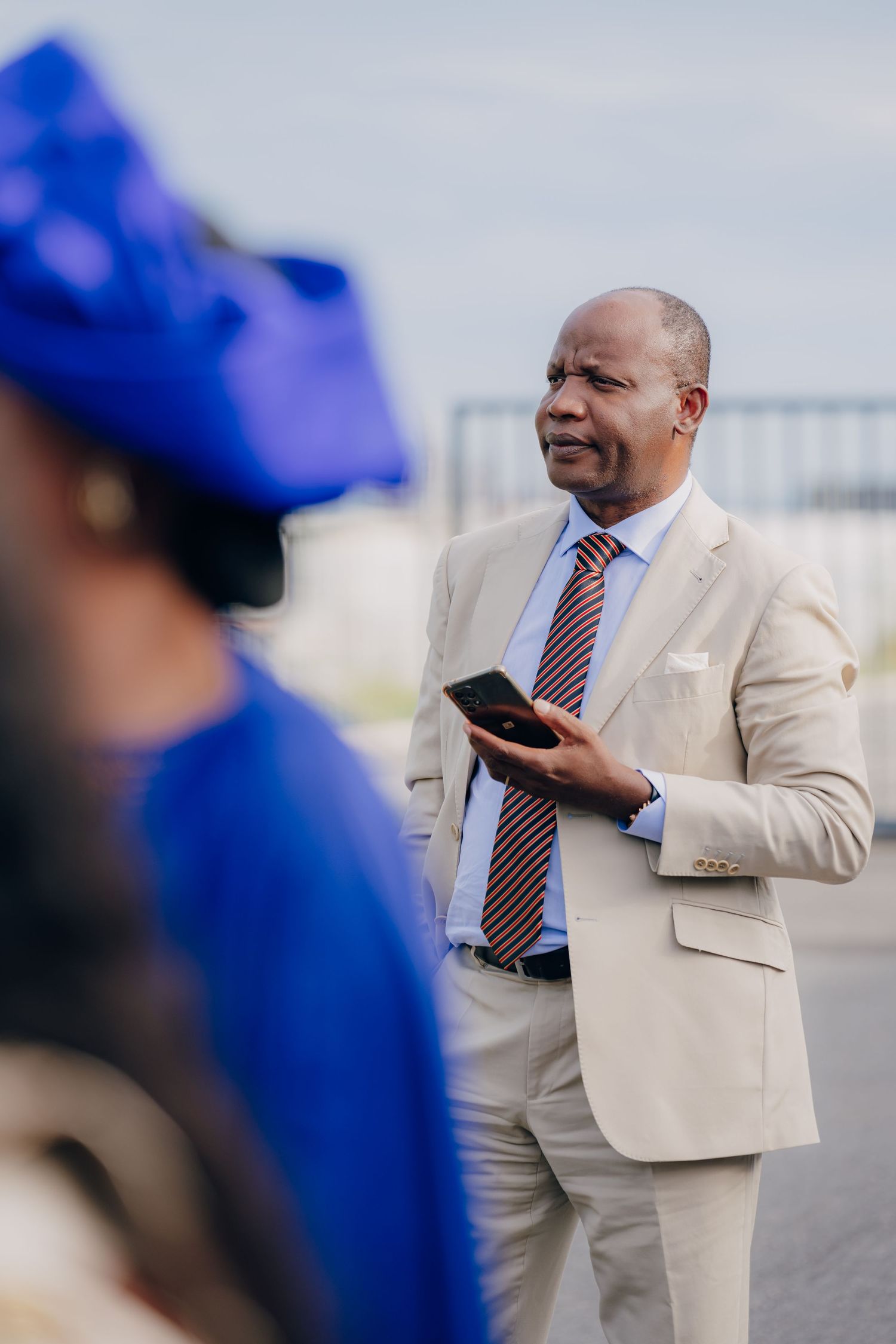 A person in a beige suit holds a phone while standing outdoors with others in blue attire visible.
