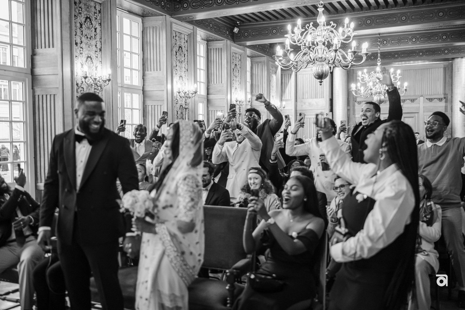 Wedding guests celebrate joyfully with raised arms in an elegant ballroom with chandeliers and ornate windows.