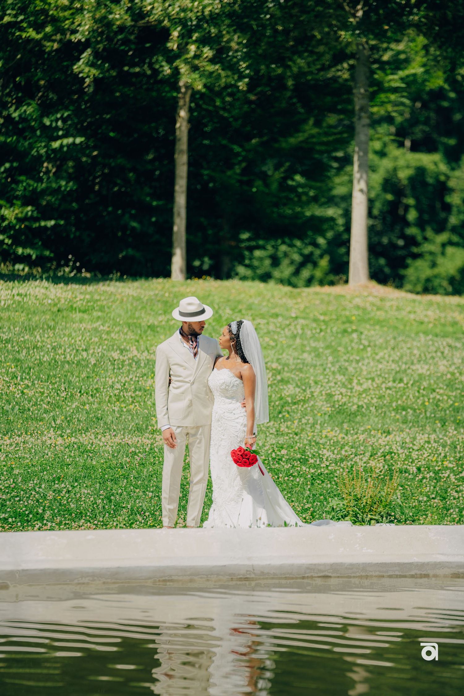 A couple in elegant wedding attire poses near a reflective pond in a lush garden setting.