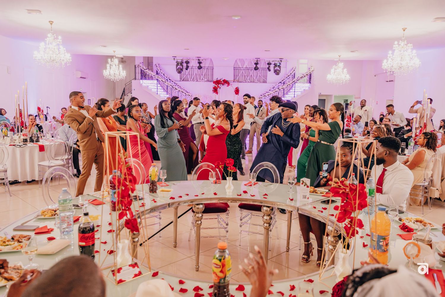 Wedding reception celebration with guests dancing under crystal chandeliers in a pink-lit ballroom.