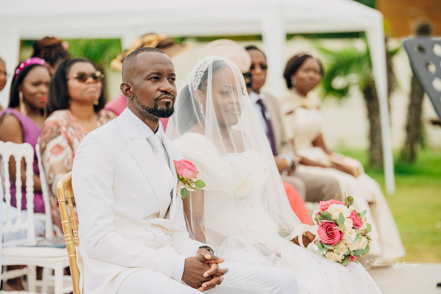 Couple seated during outdoor garden wedding ceremony under white tent.