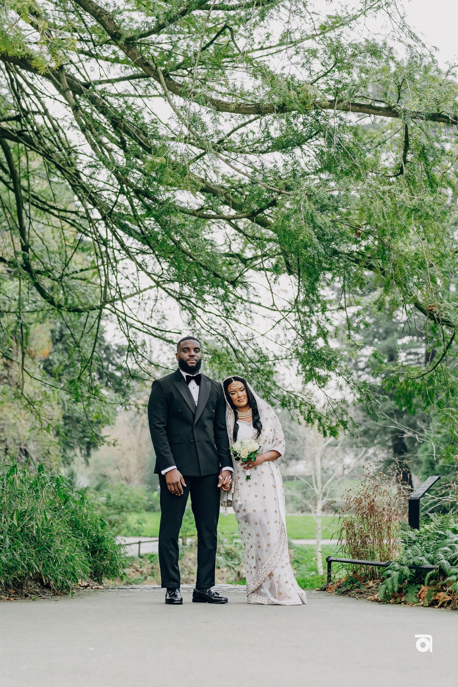 A couple in wedding attire stands on a path under overhanging trees and lush greenery in a garden setting.
