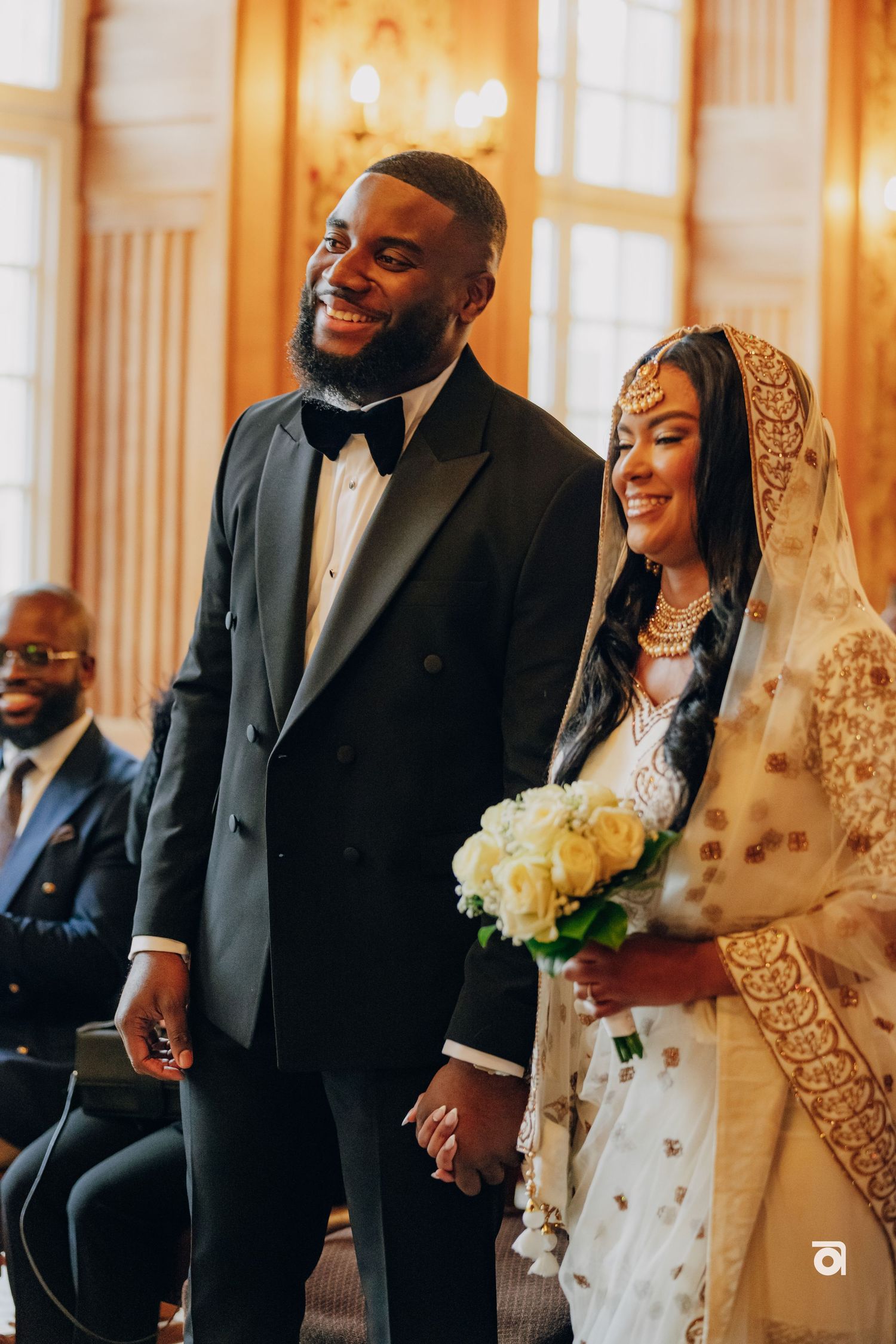 A happy wedding couple smiling in formal attire with the bride in a white and gold veil holding yellow roses.