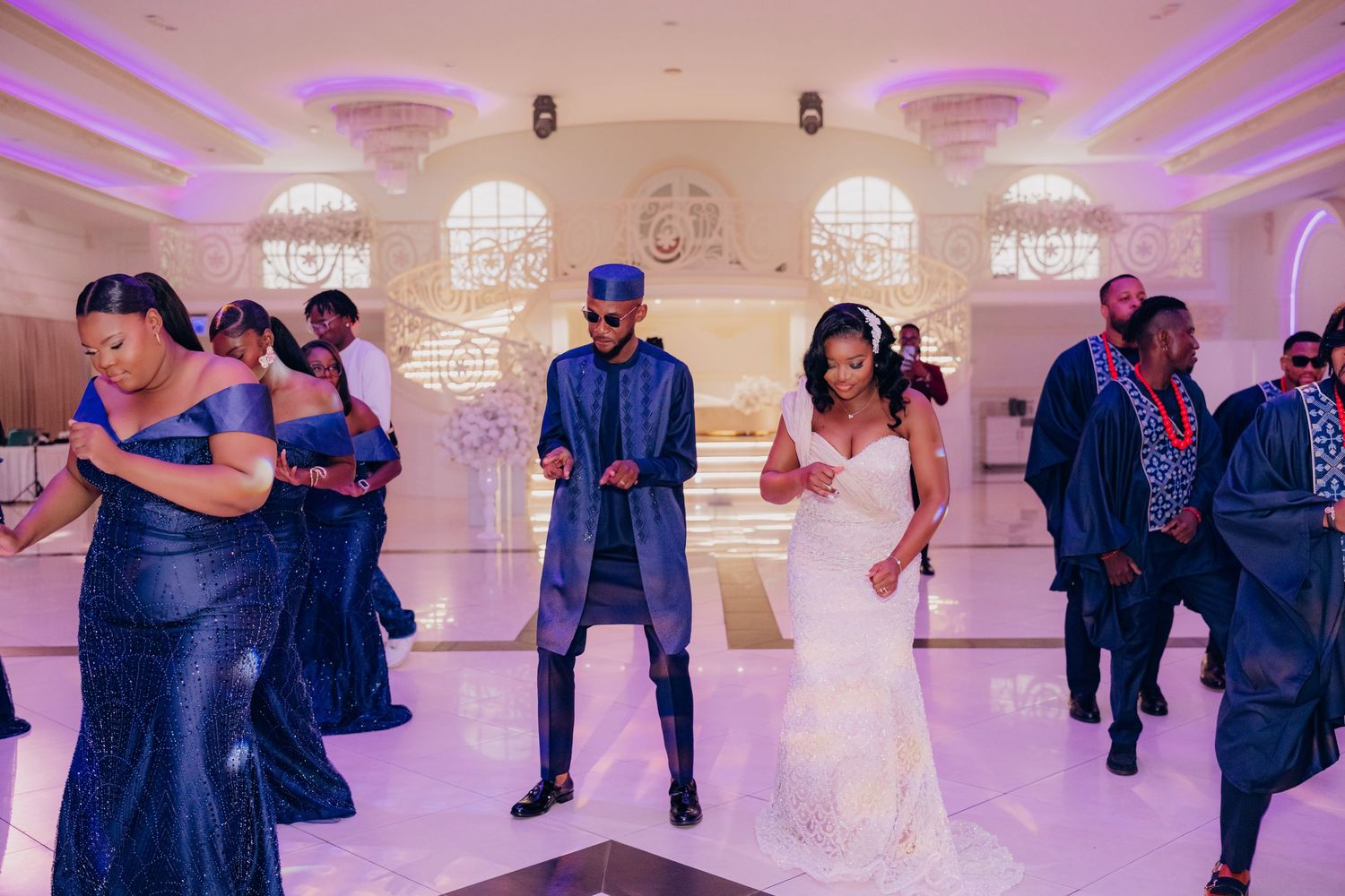 Wedding guests dance together on an elegant ballroom floor illuminated with purple lighting and crystal chandeliers.