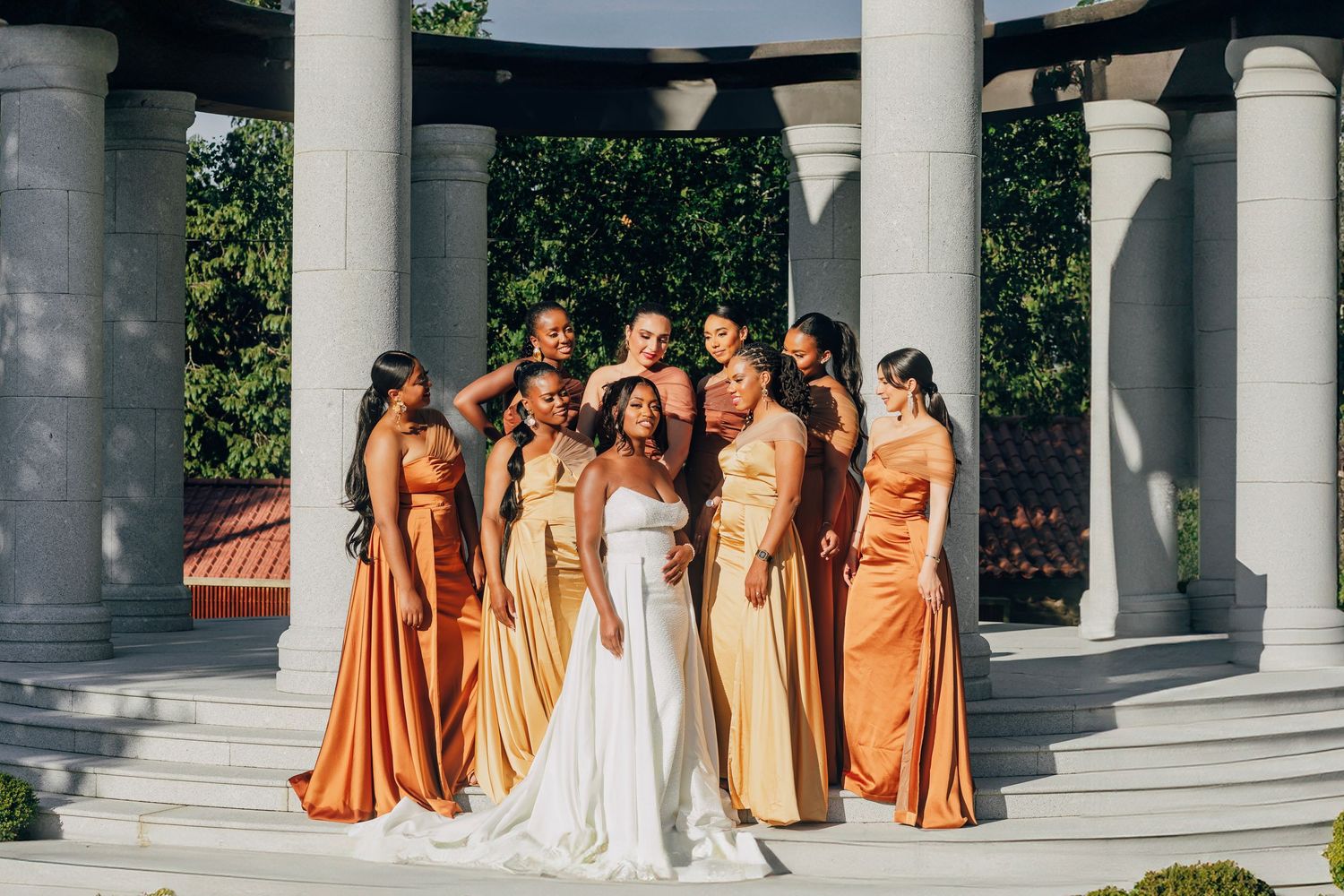 A bridal party poses in gold and orange dresses under a columned pavilion.