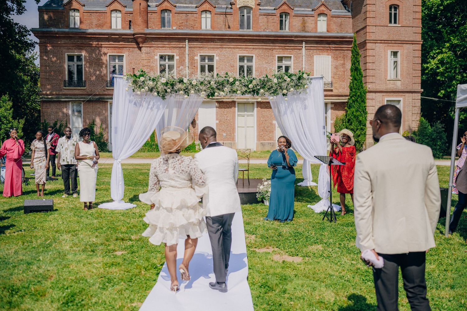 Wedding guests gather on a lush green lawn outside a grand historic brick mansion with ornate architectural details.