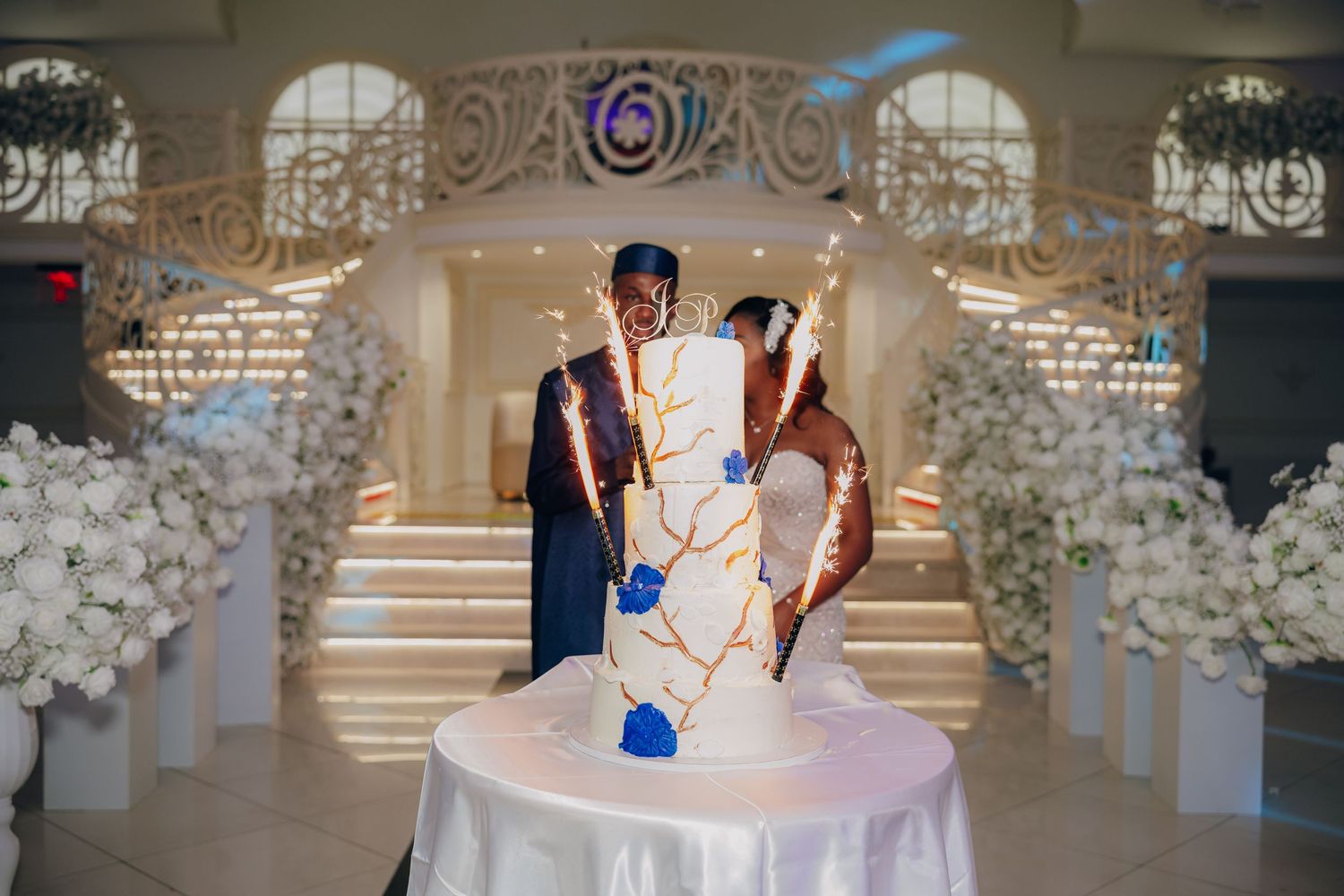 Wedding cake topper displayed on decorated table with ornate floral arch and staircase in background.