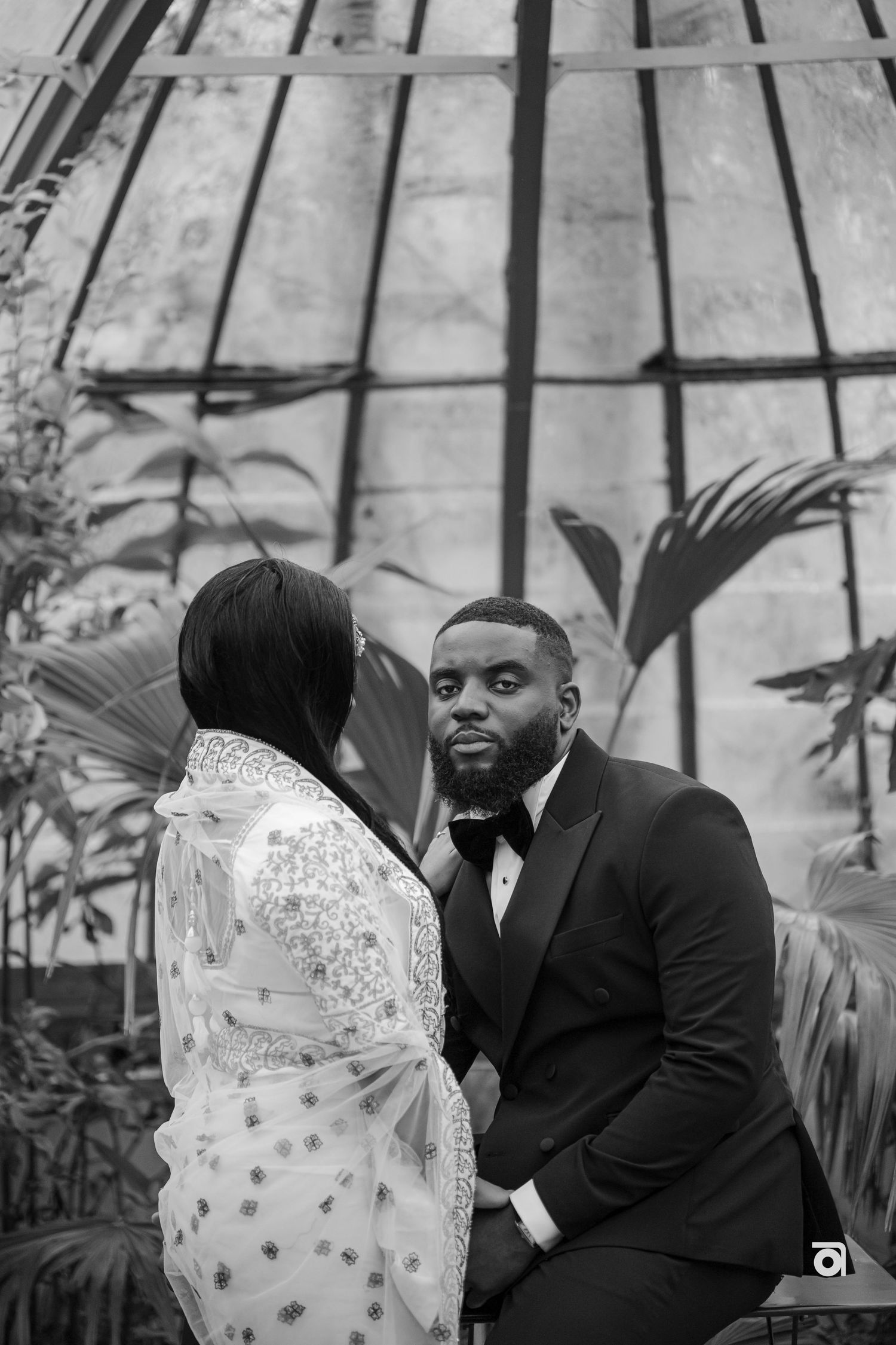 Black and white wedding photo captured inside an elegant greenhouse with palm fronds in background.