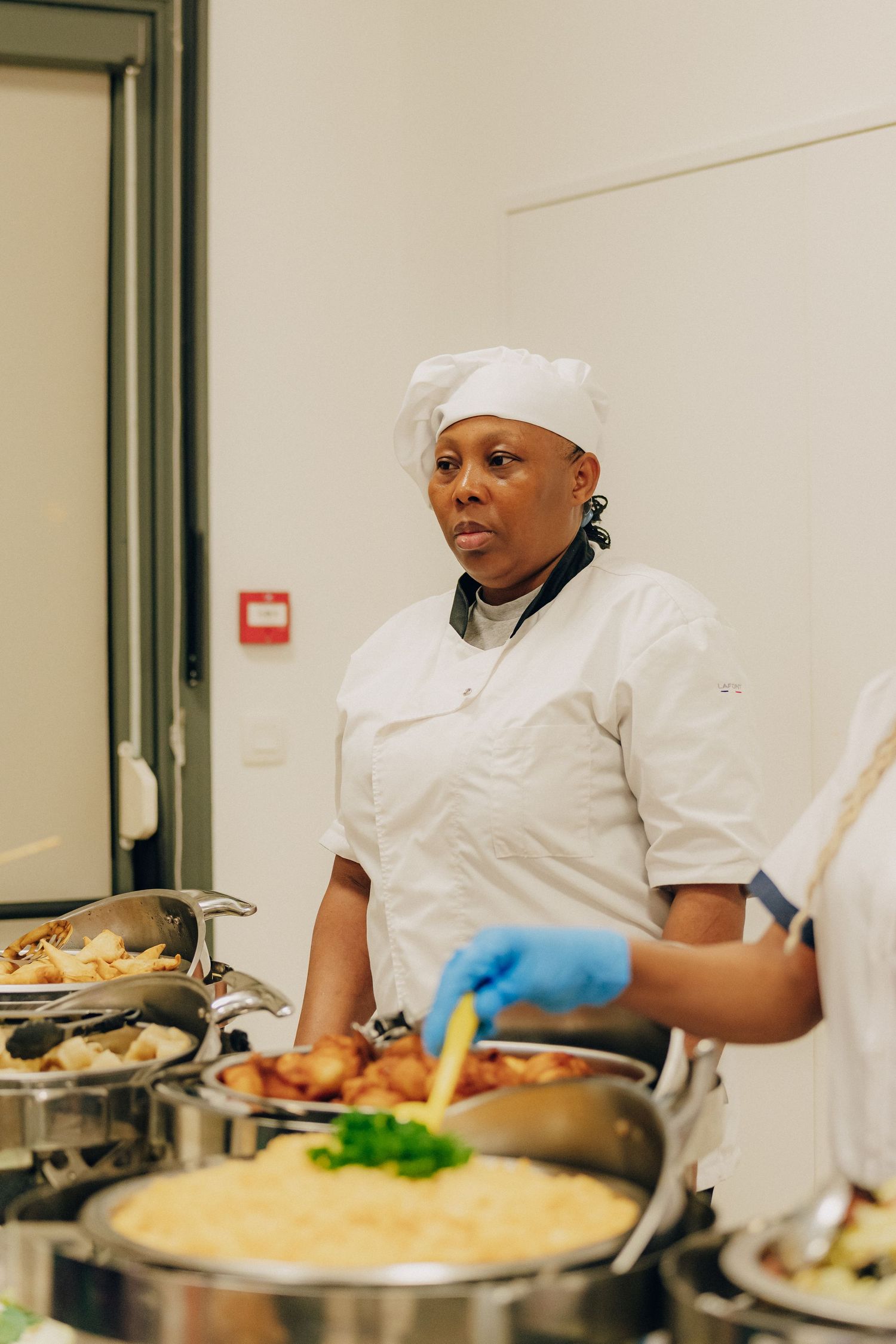 Chef in white uniform and head covering stands at catering station with prepared dishes.