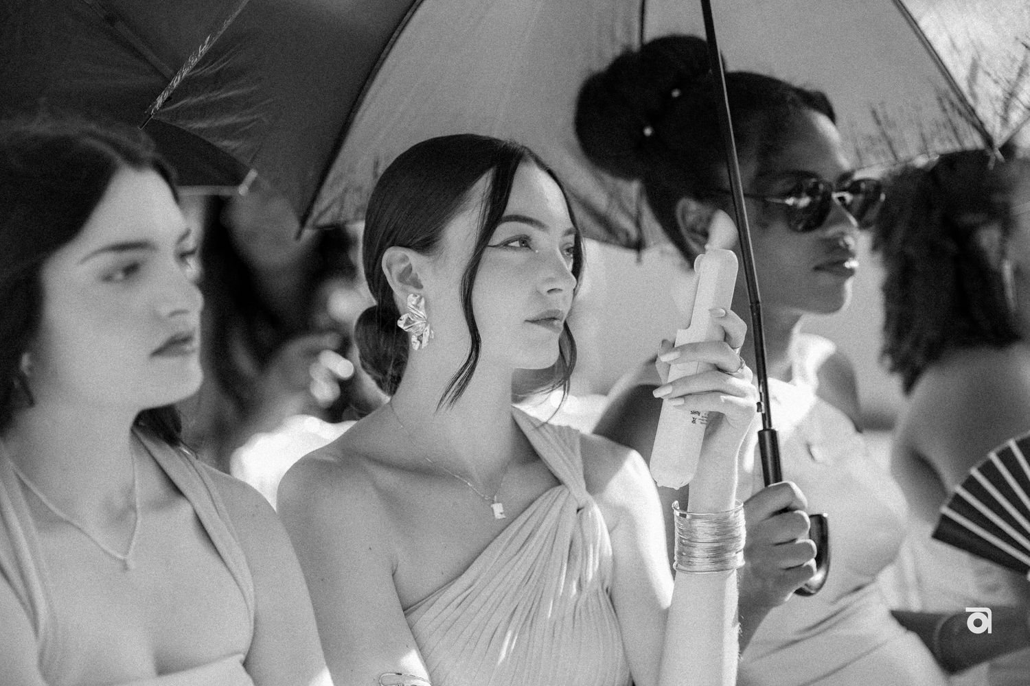 A black and white photo of guests holding umbrellas and fans during an outdoor event.