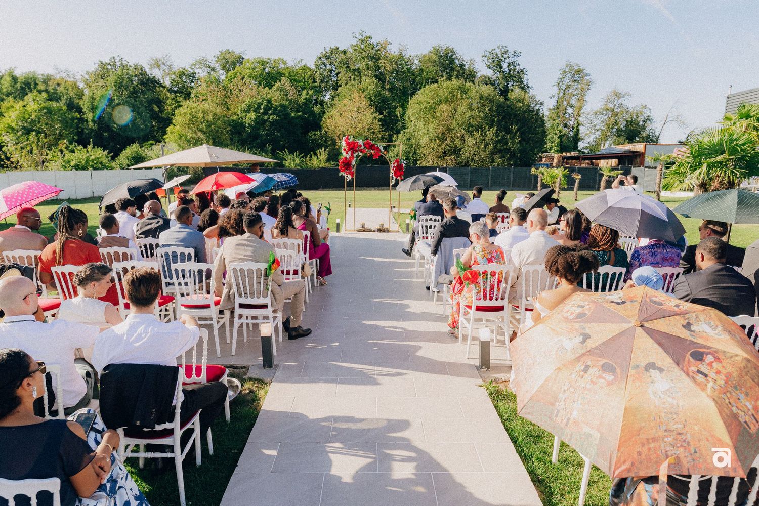 Outdoor wedding ceremony setup with rows of white chairs and guests holding decorative parasols on a sunny day.