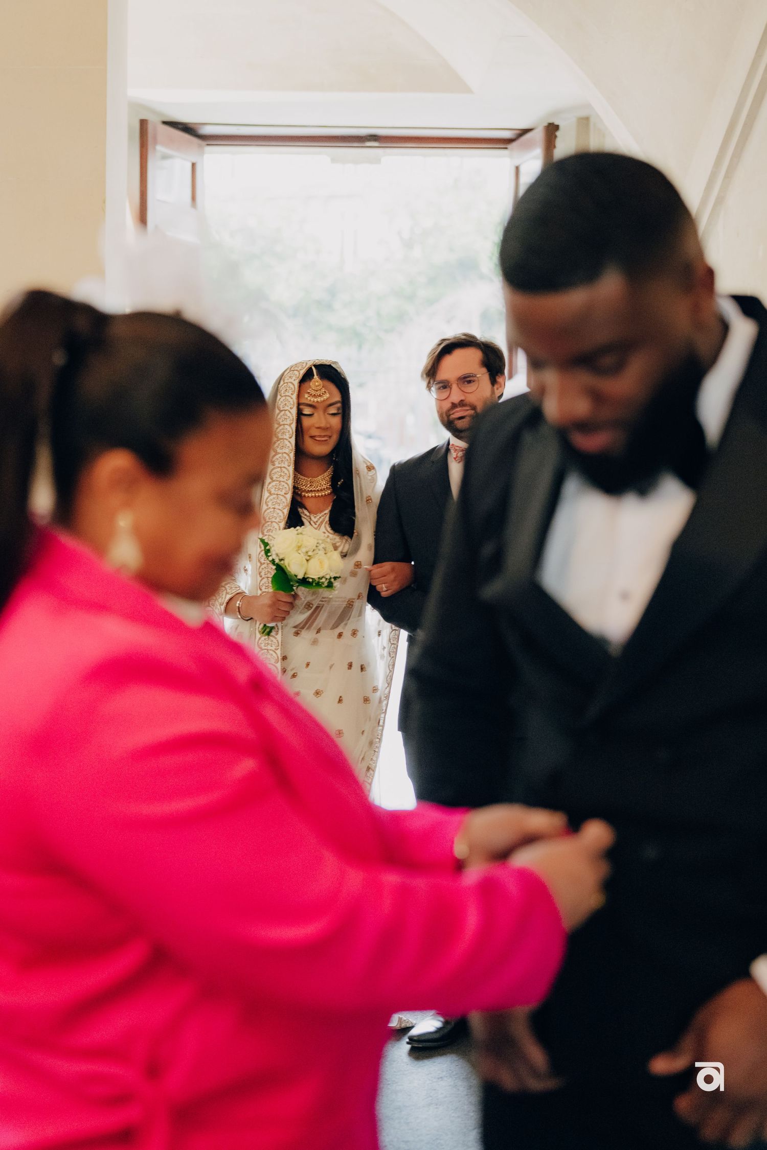 Wedding party members gather in doorway as bride in embellished gown stands with bouquet.