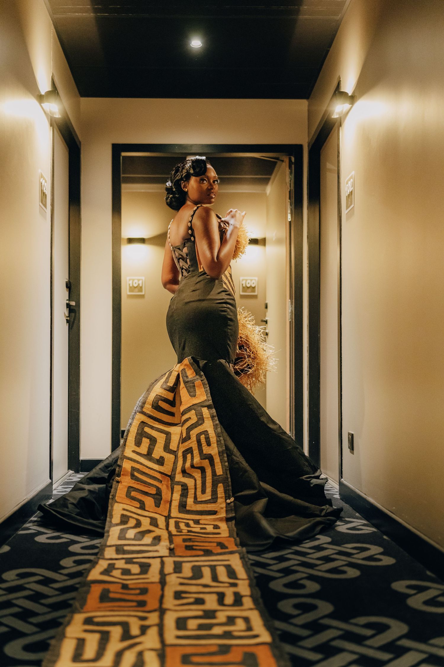 A model in a dark mermaid-style evening gown poses in a hotel hallway with geometric patterned carpet.
