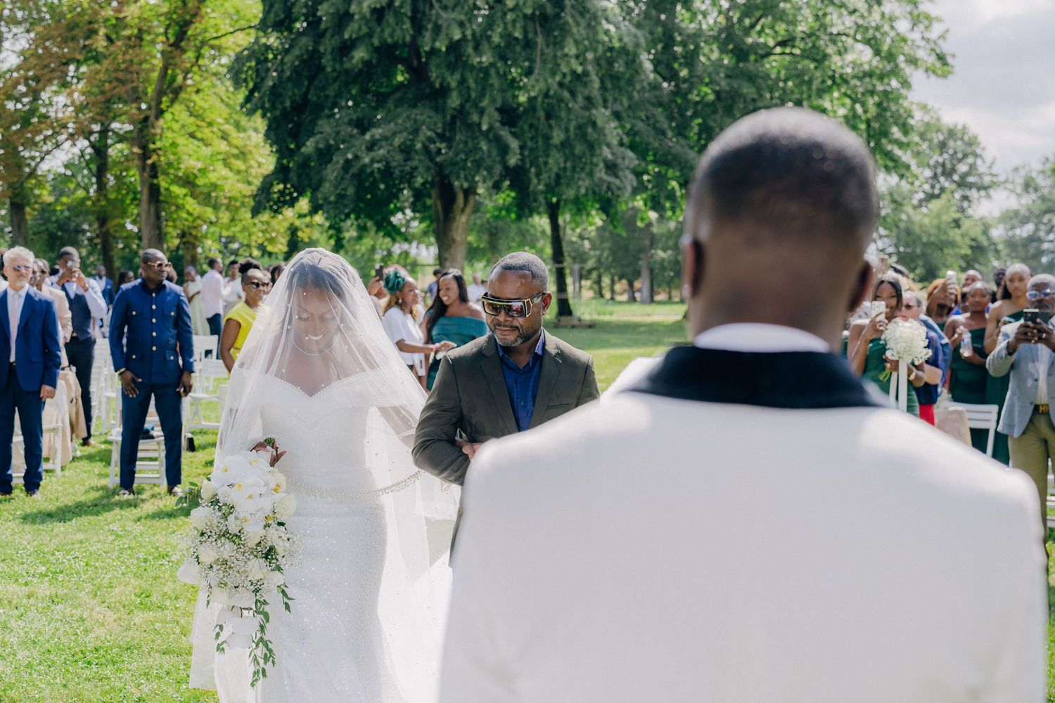 Wedding party walking through a lush green garden path with guests looking on during a summer celebration.