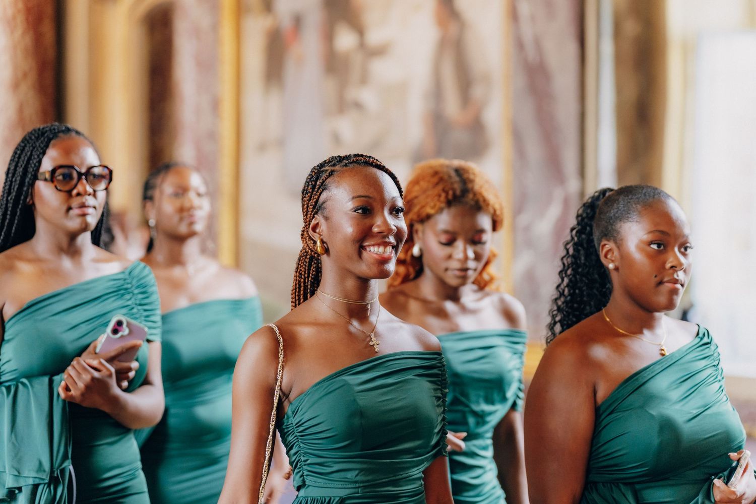Bridesmaids in matching teal one-shoulder dresses share joyful reactions during wedding celebration.