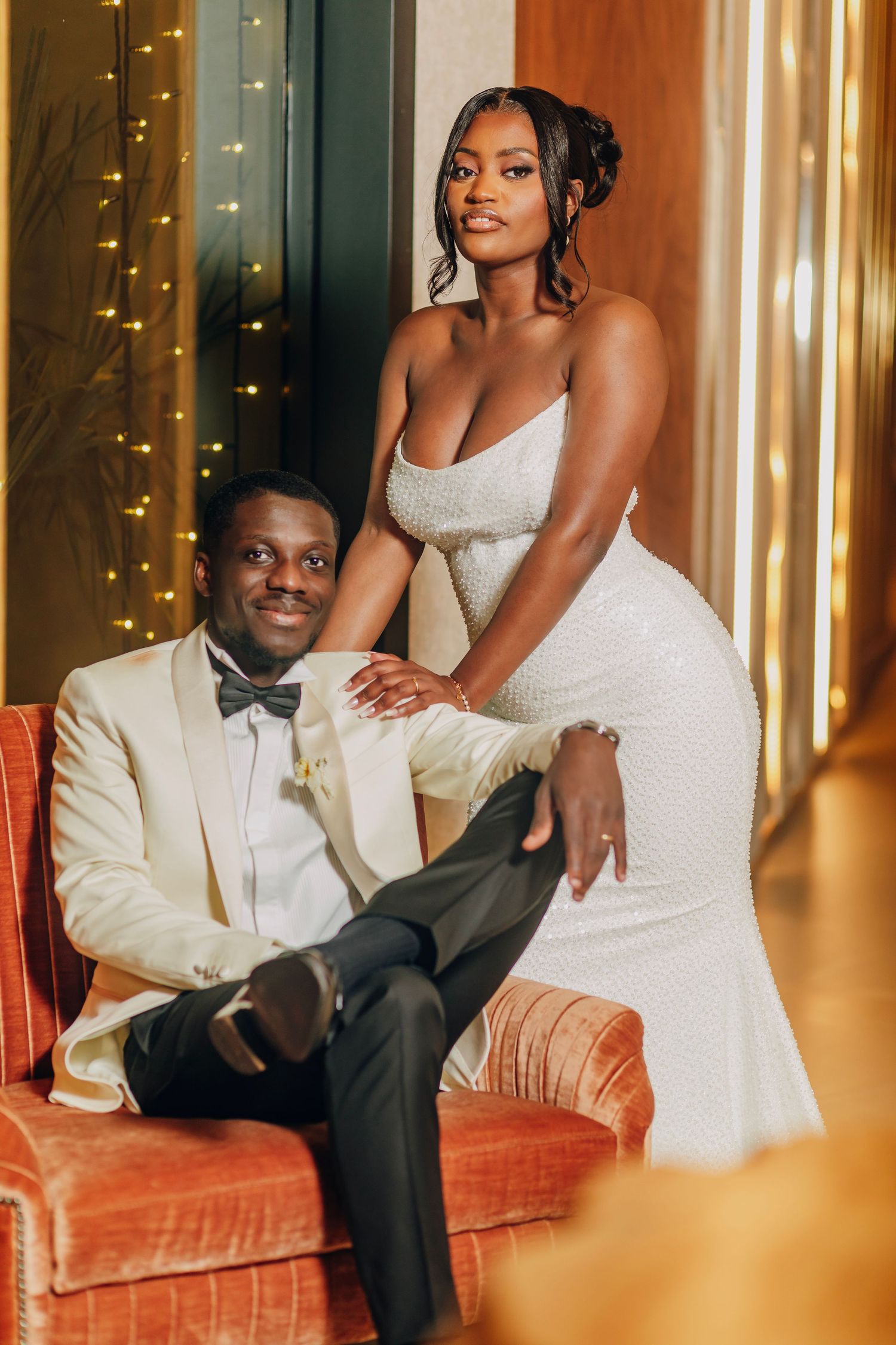Elegantly dressed couple poses in formal attire against sparkling fairy light backdrop.