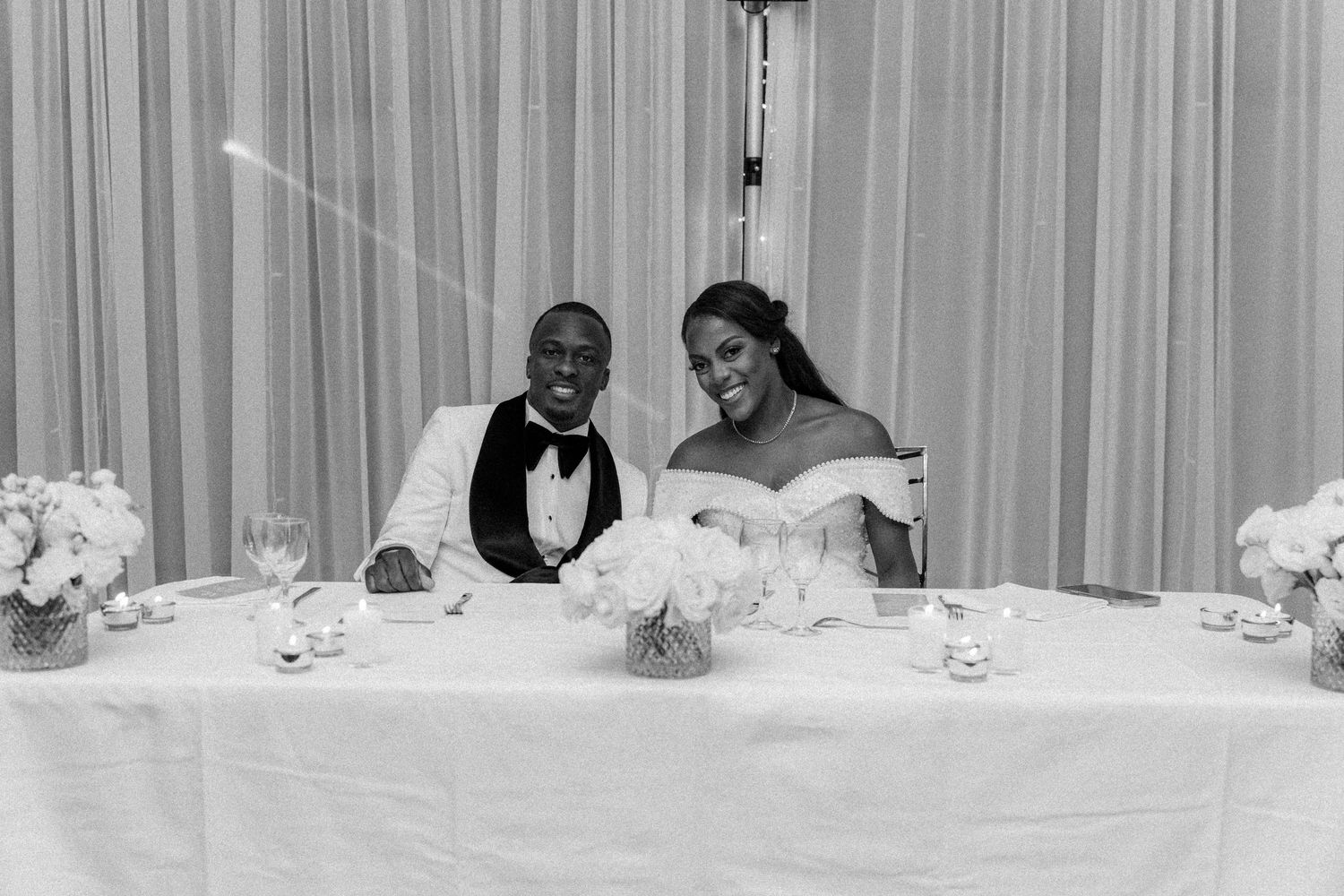 Elegant white wedding reception head table setup with floral centerpieces and draped backdrop.