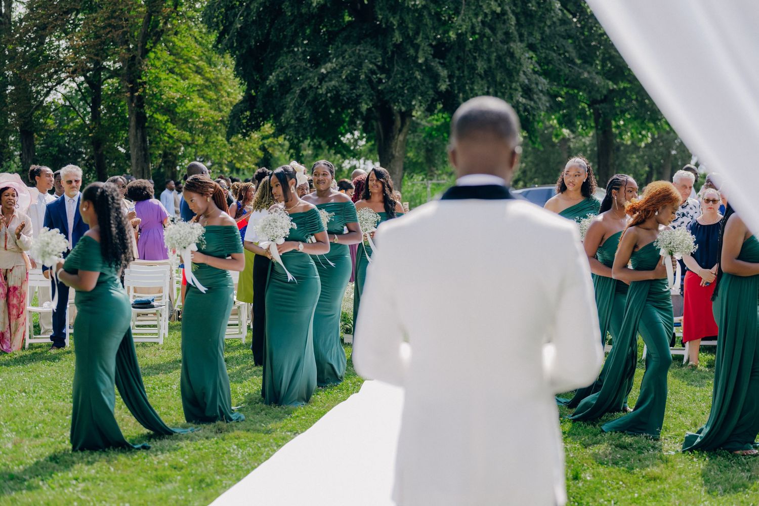 Wedding party members in emerald green dresses gather on a sunny lawn during an outdoor ceremony.
