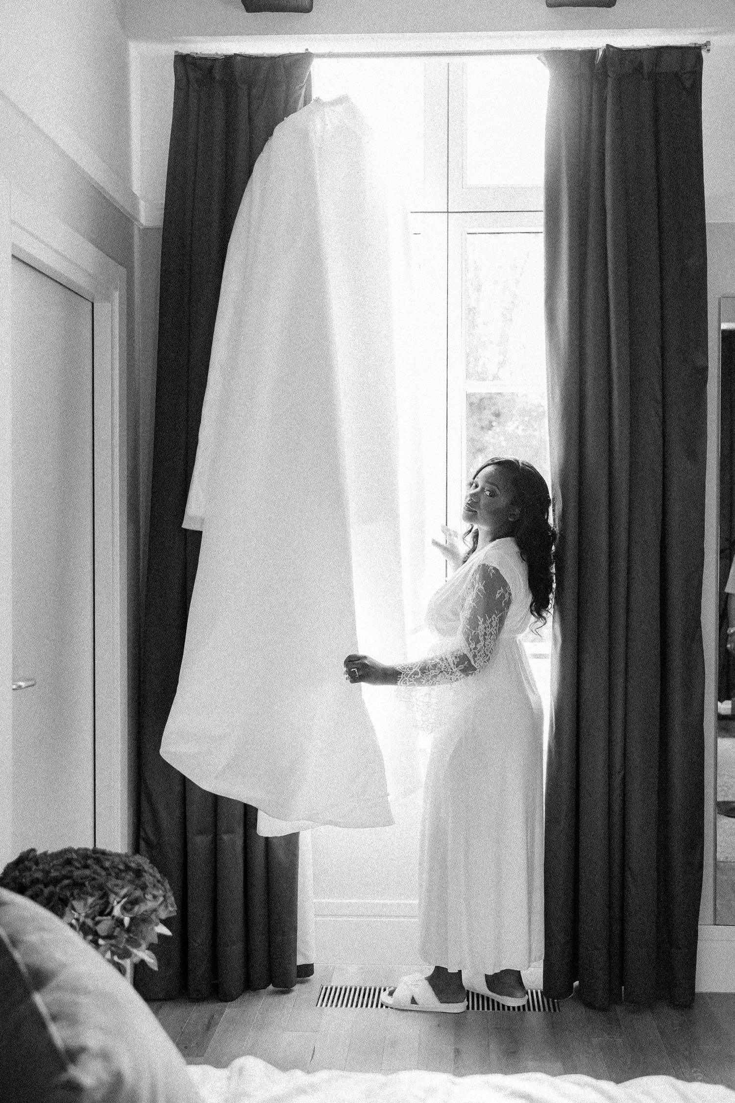 White wedding dress hanging in sunlit room with dark curtains and window.