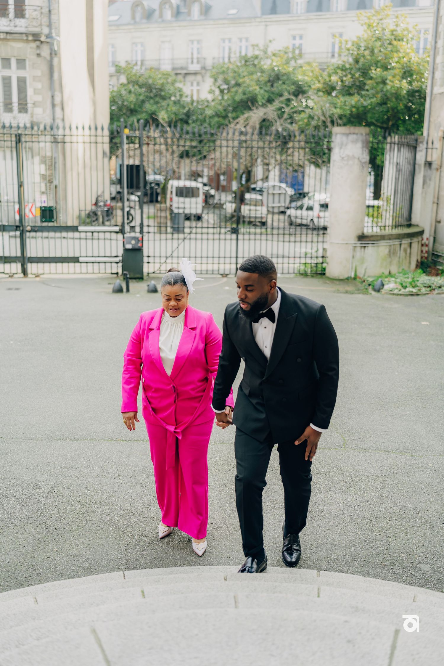 A couple in contrasting formal wear - hot pink suit and black tuxedo - walk together on stone steps.