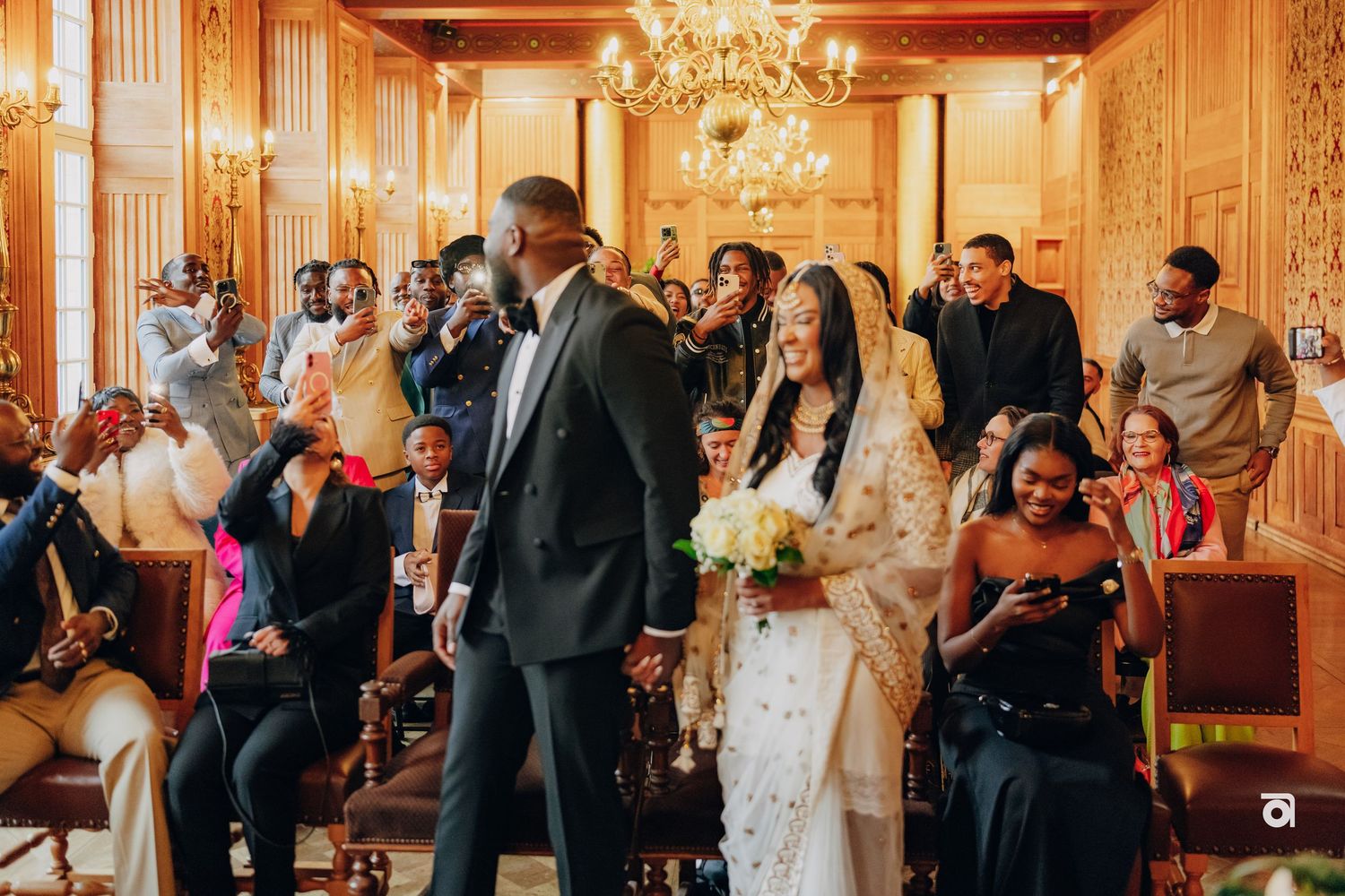 Wedding guests celebrate in an elegantly decorated room with chandelier and wooden beams.