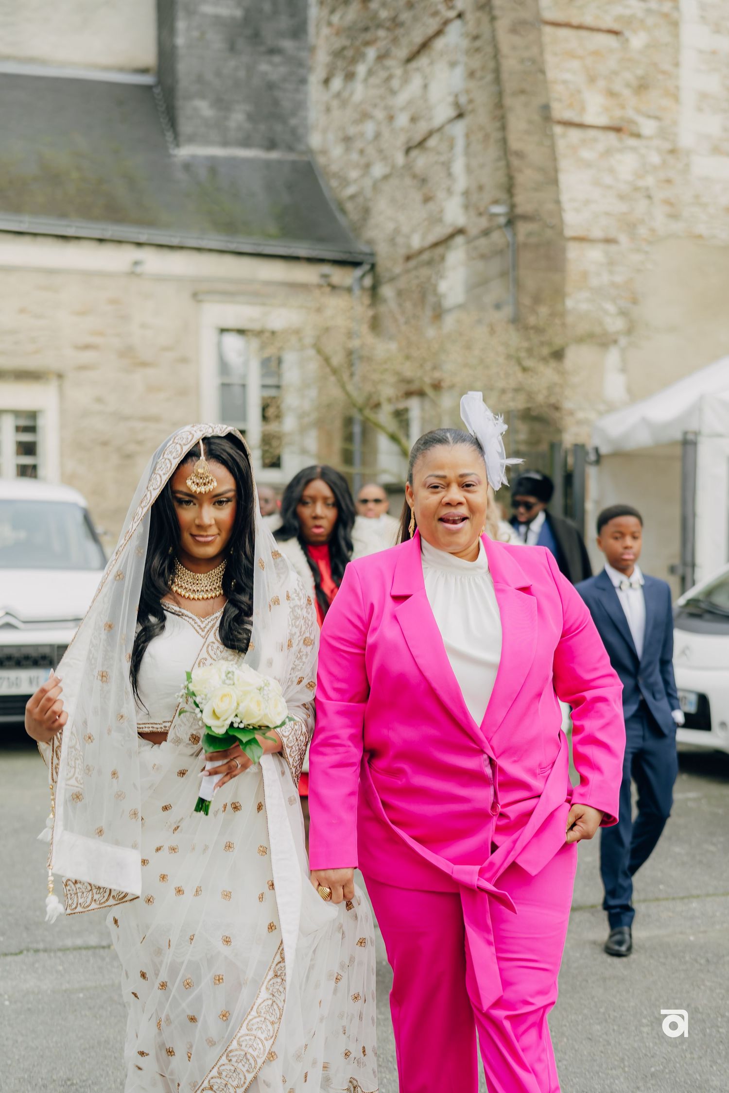 Bride in white embellished dress walks alongside person in bright pink suit at wedding entrance.
