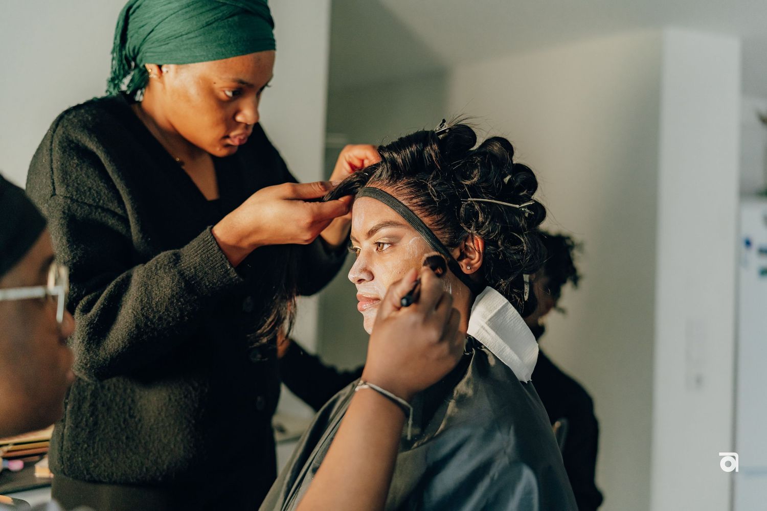 A hairstylist wearing a green head wrap works carefully on styling curly hair in a bright salon setting.
