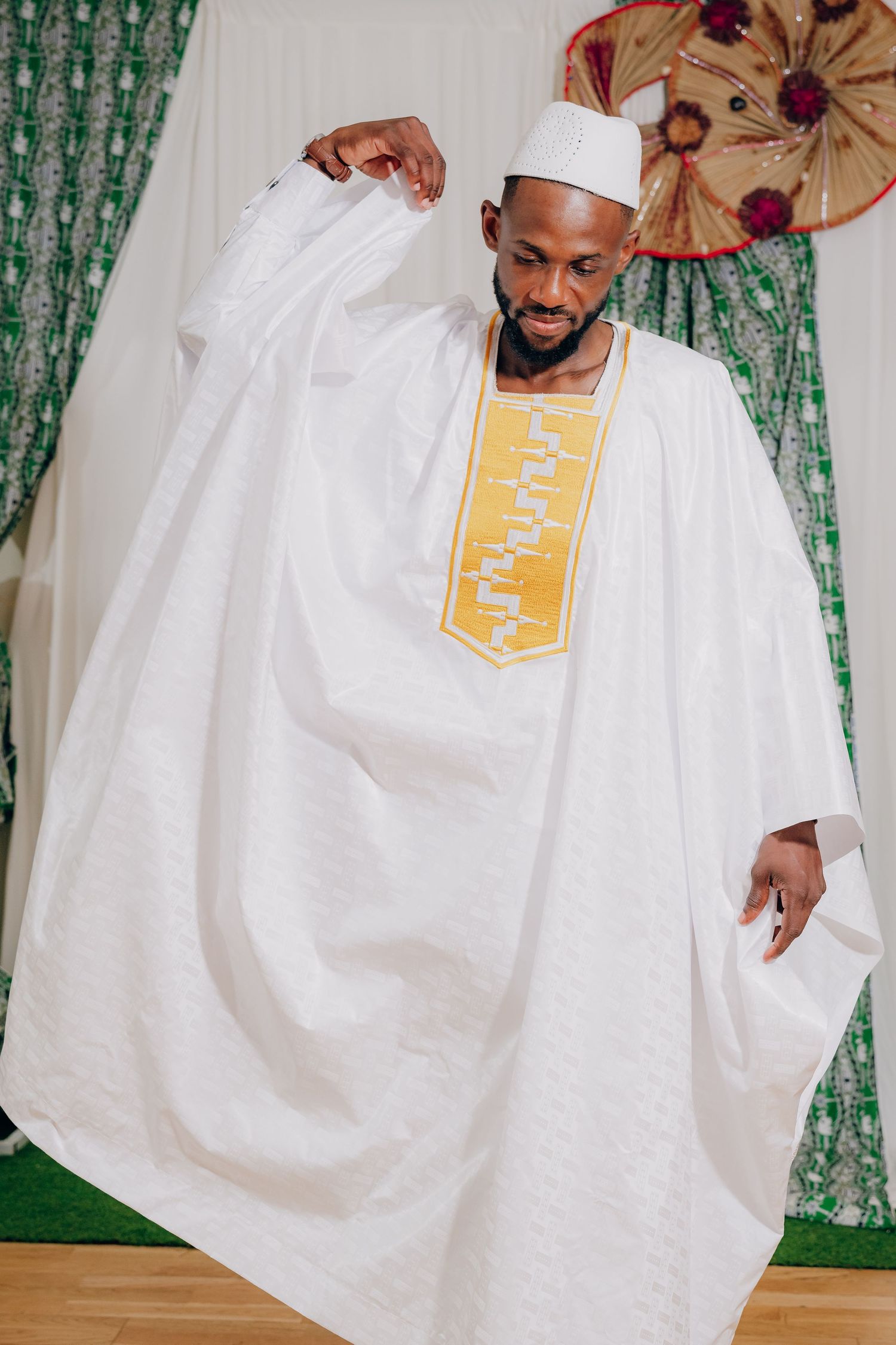 A person wearing a flowing white traditional agbada robe with red and gold accents poses against a green backdrop.