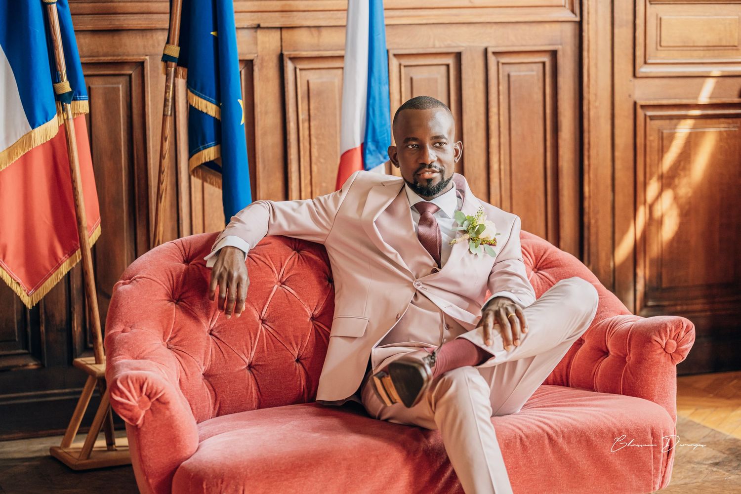 Elegant pink suit-clad figure poses on vintage red velvet couch with flags in background.