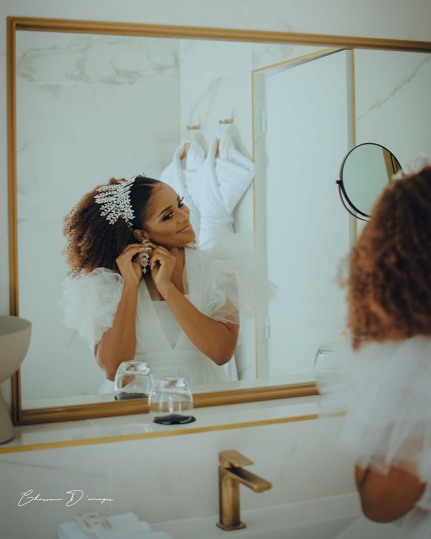 A bride puts on jewelry while looking in a bathroom mirror with a white dress hanging in background.