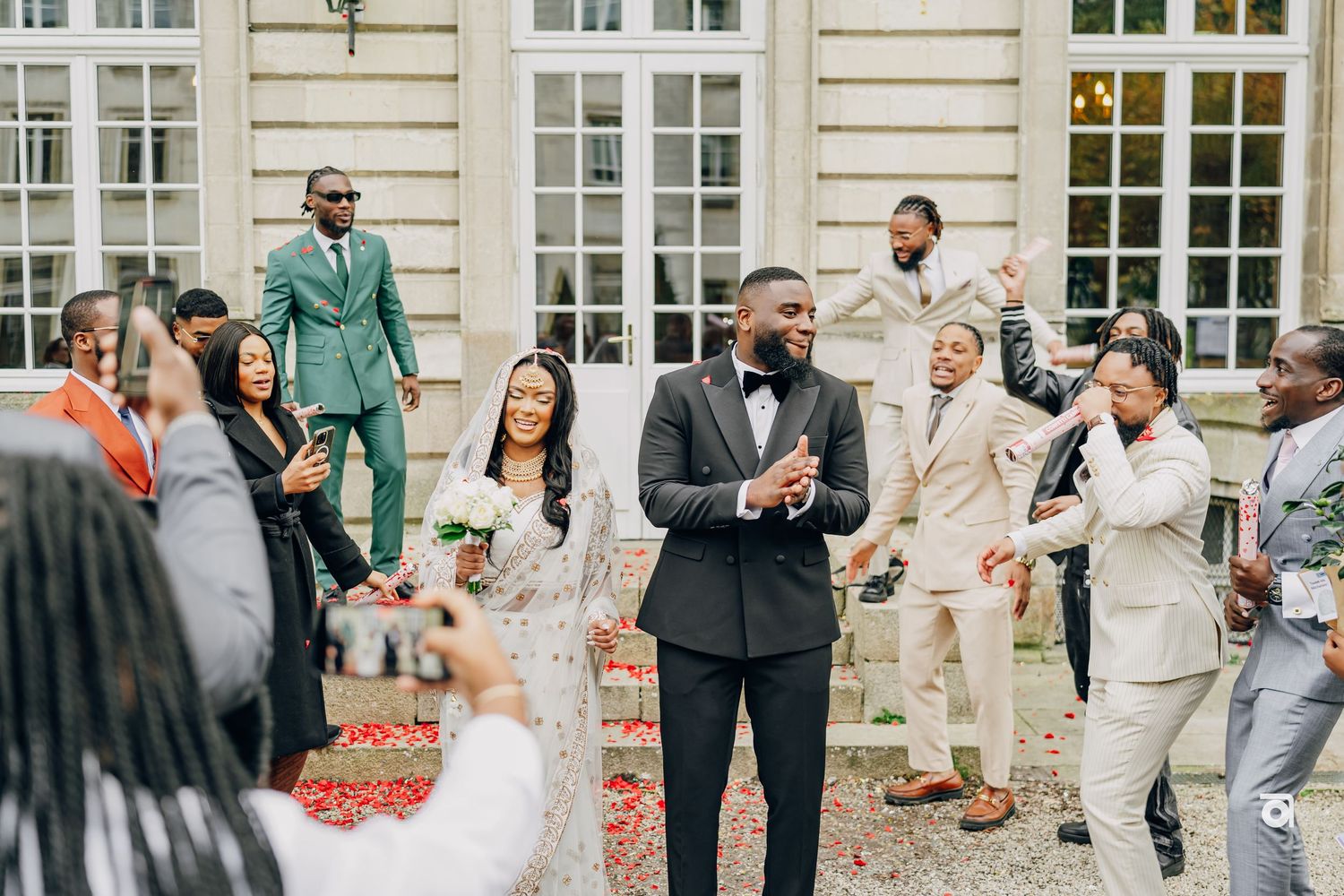 Wedding party celebrates outside elegant white building with guests in pastel suits.