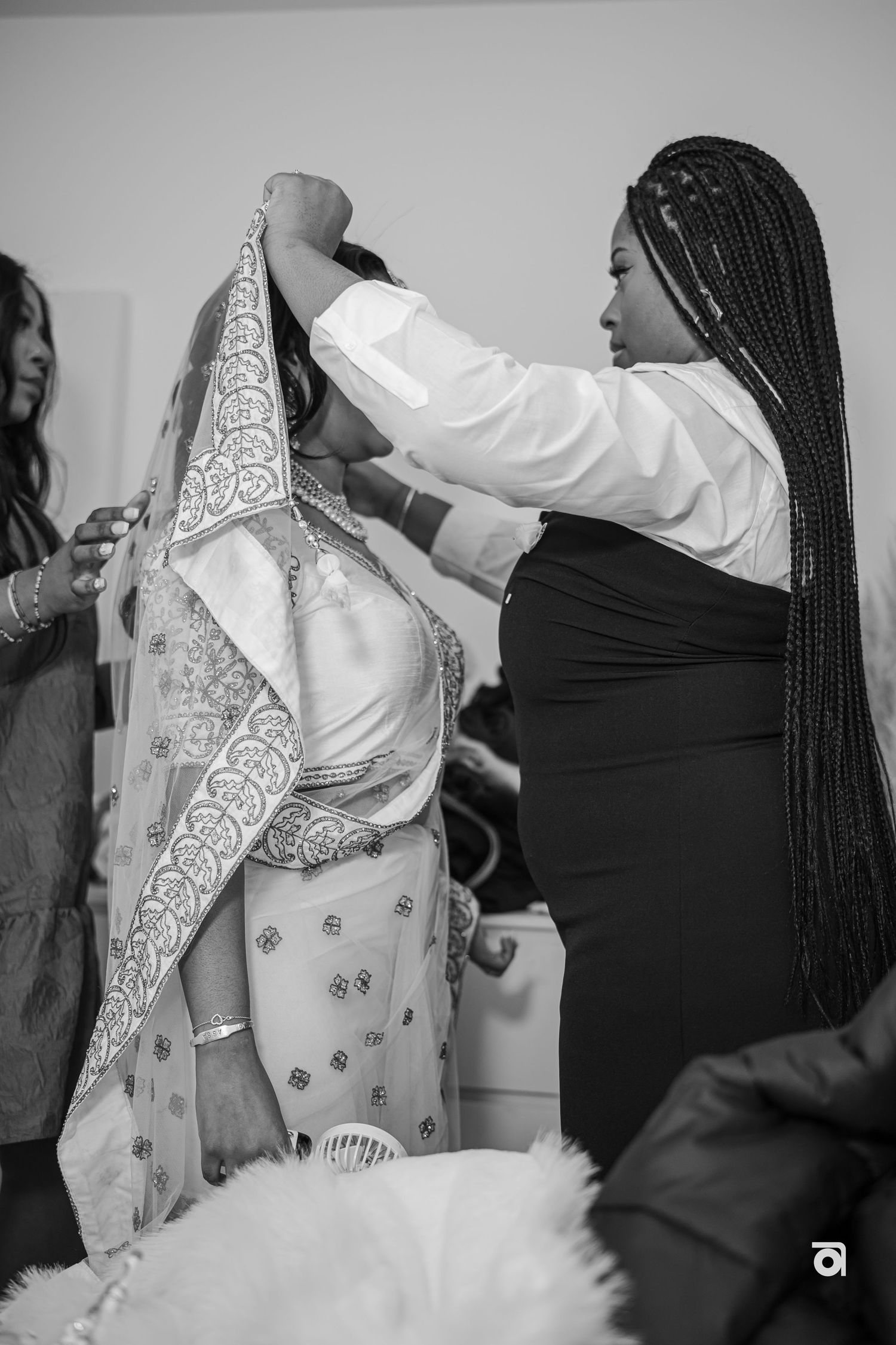 A person in black helps adjust traditional wedding attire with intricate lace details during preparation.