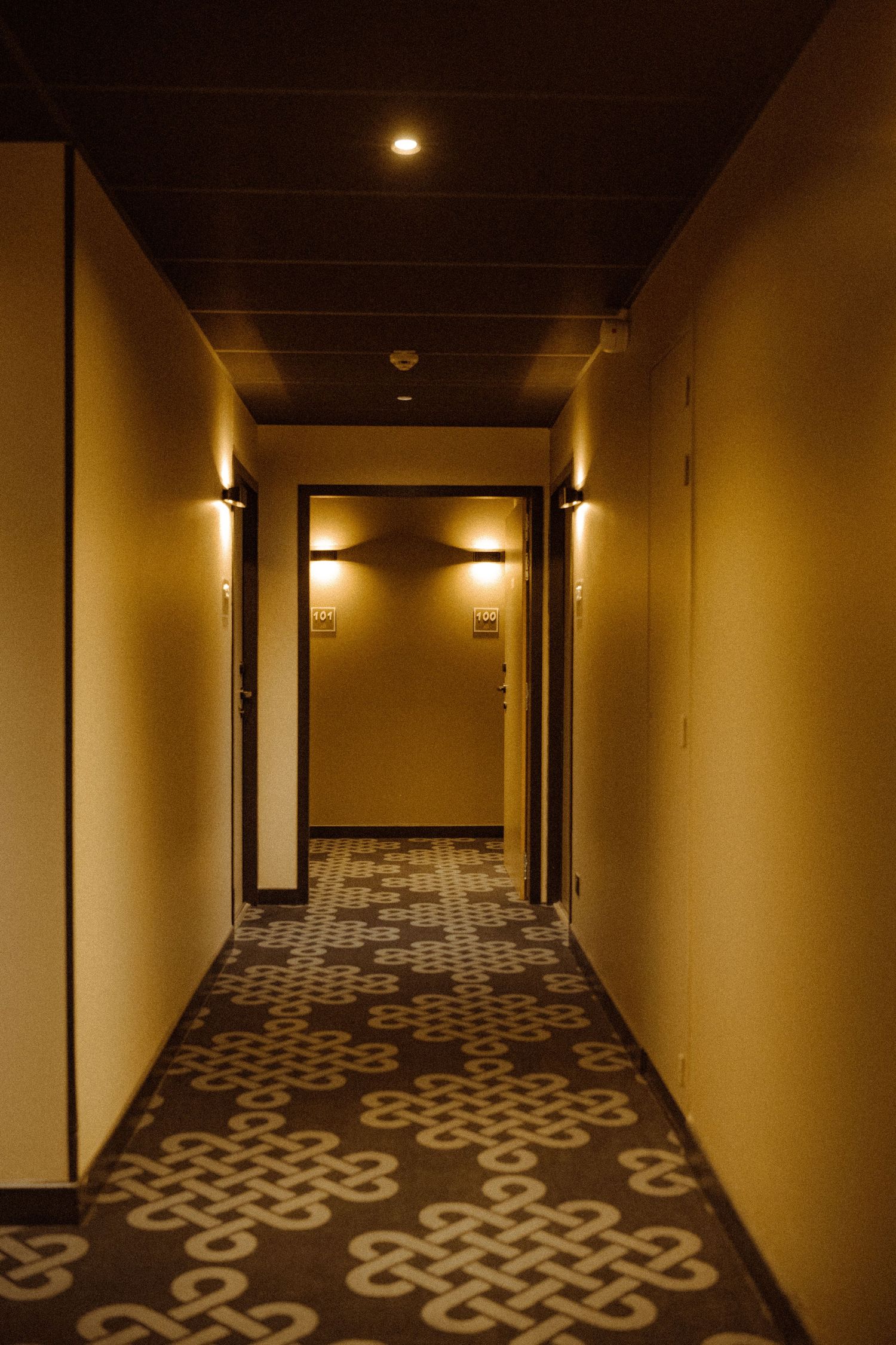 A dimly lit hotel hallway with patterned carpet and warm wall sconces creating an atmospheric glow along the corridor.