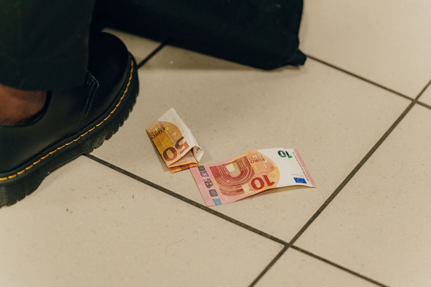 Euro banknotes lying on white tiled floor next to black boot.