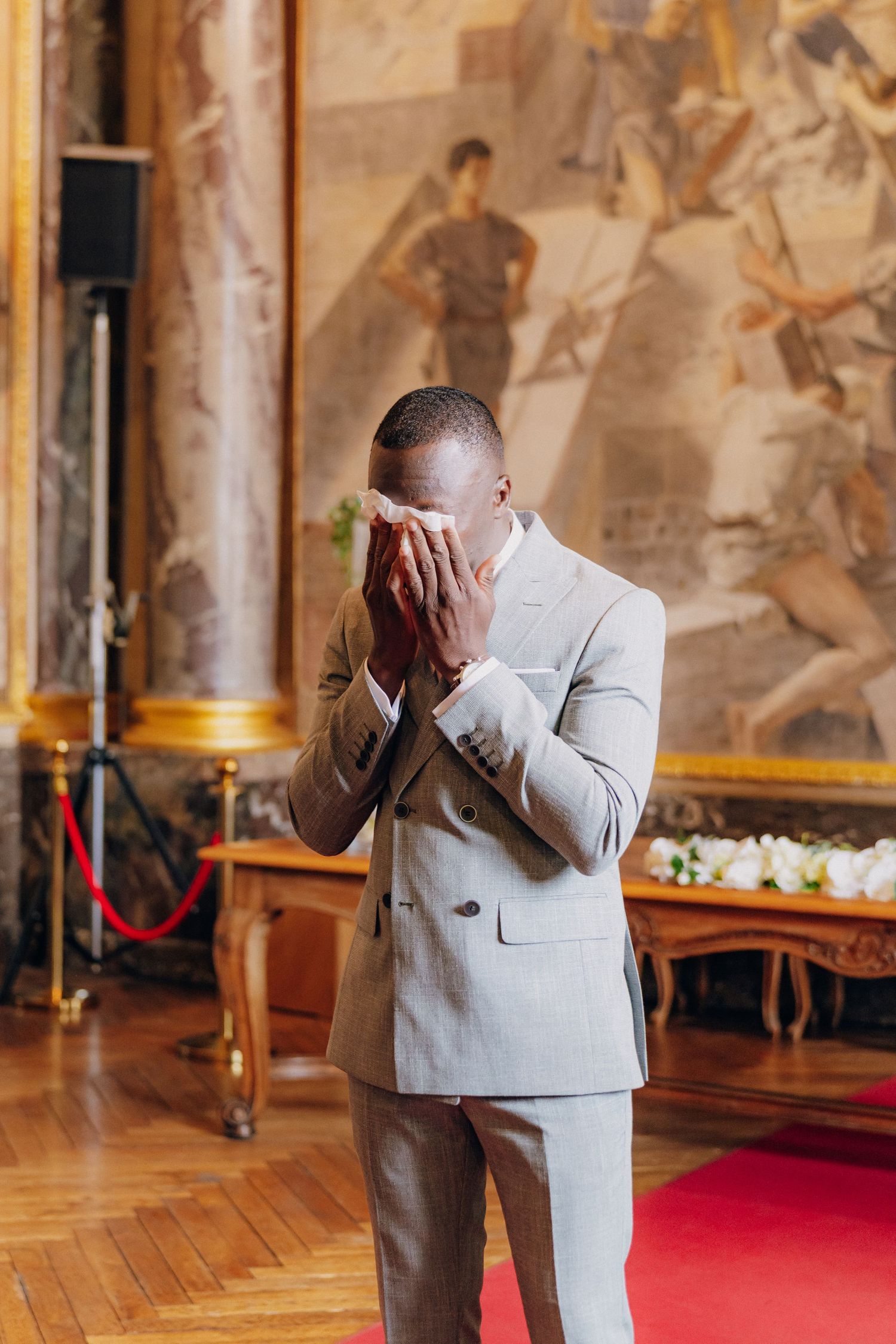 A person in a gray suit covers their face with both hands while standing in a grand gallery.