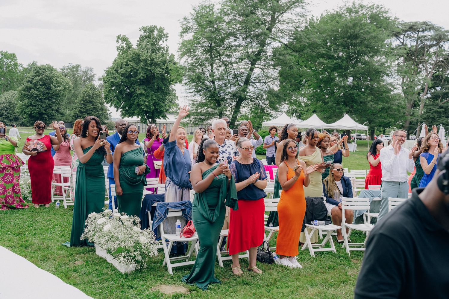 Wide shot of wedding guests seated in rows of white chairs during an outdoor ceremony.