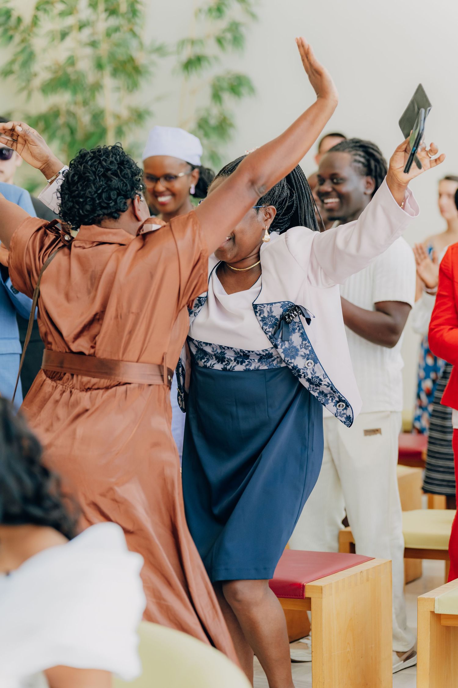 A joyful group of people dancing and celebrating together at a gathering with greenery in the background.