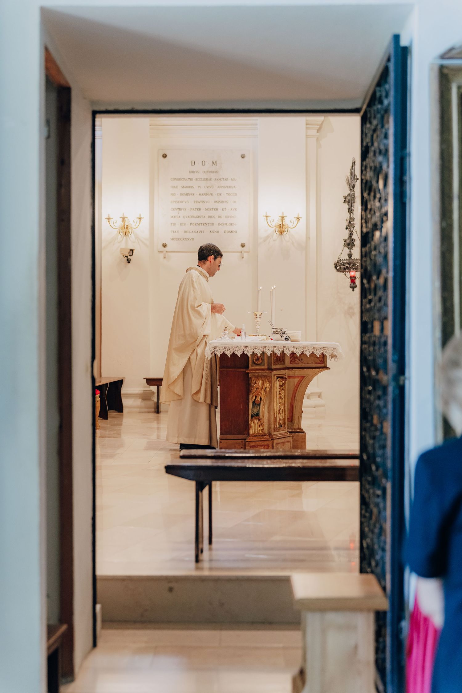 A priest in white robes conducts a religious ceremony in a bright, minimalist chapel interior.