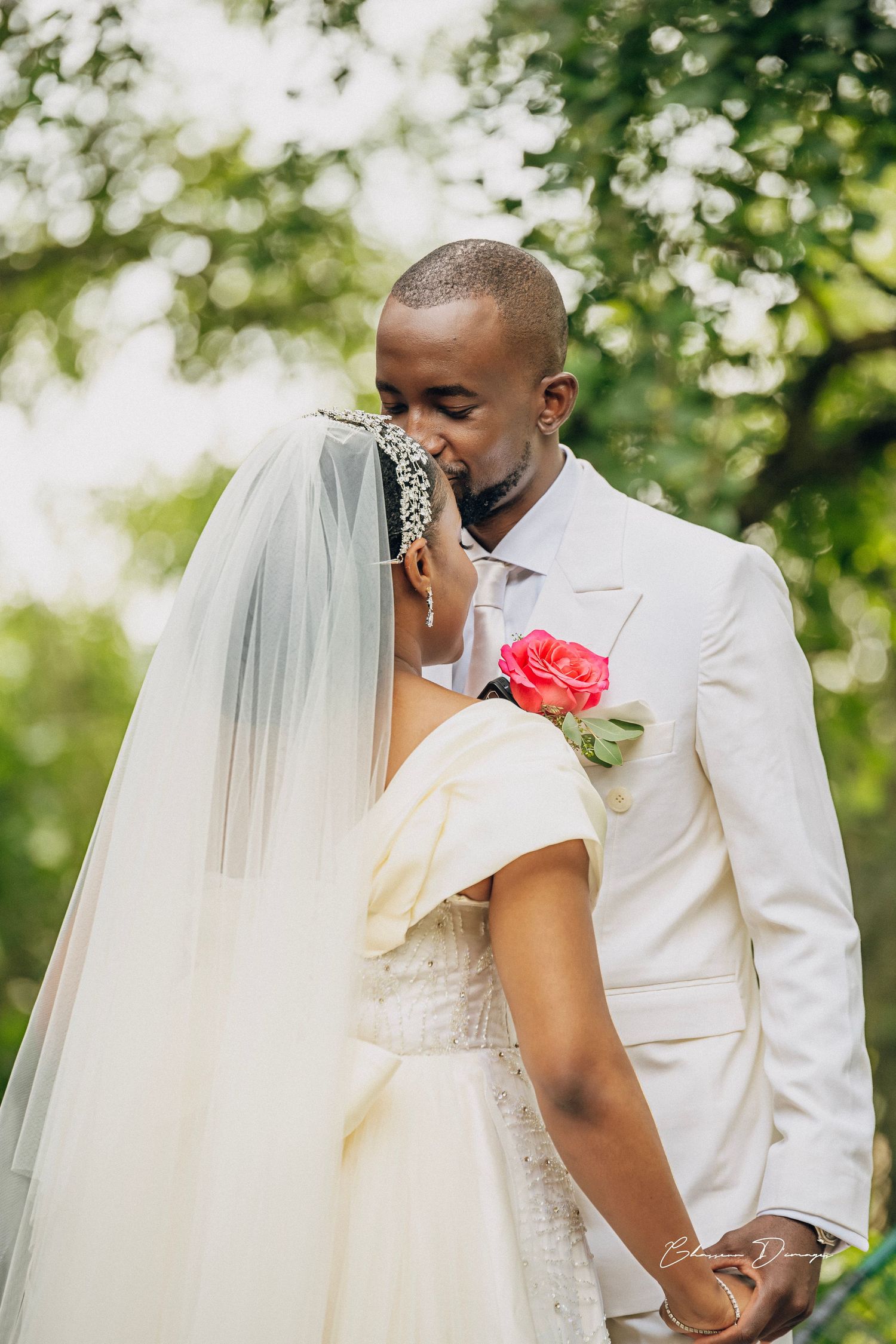 Intimate wedding moment captured under green trees with white suit and flowing veil.