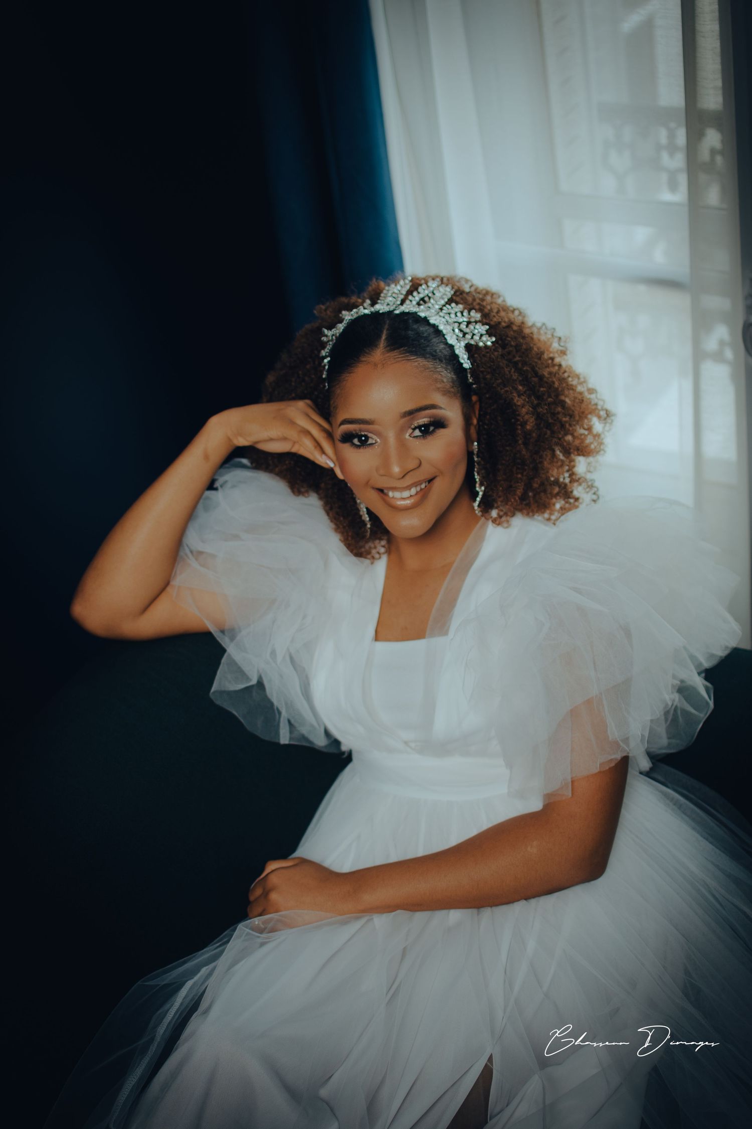 A bride wearing a white tulle dress and crystal headpiece poses by a window.