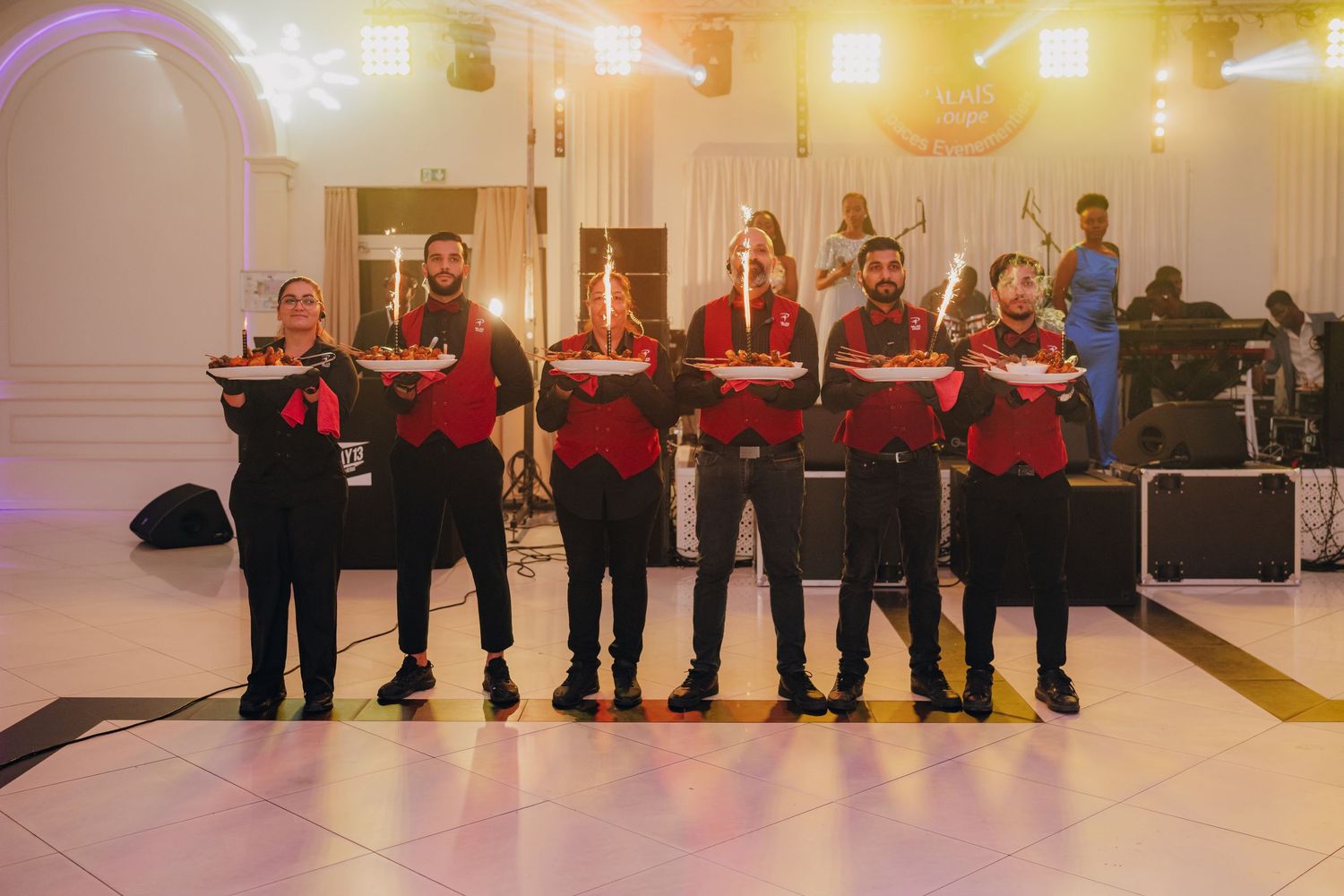 A line of musicians in red uniforms perform at an elegant indoor venue with purple lighting.