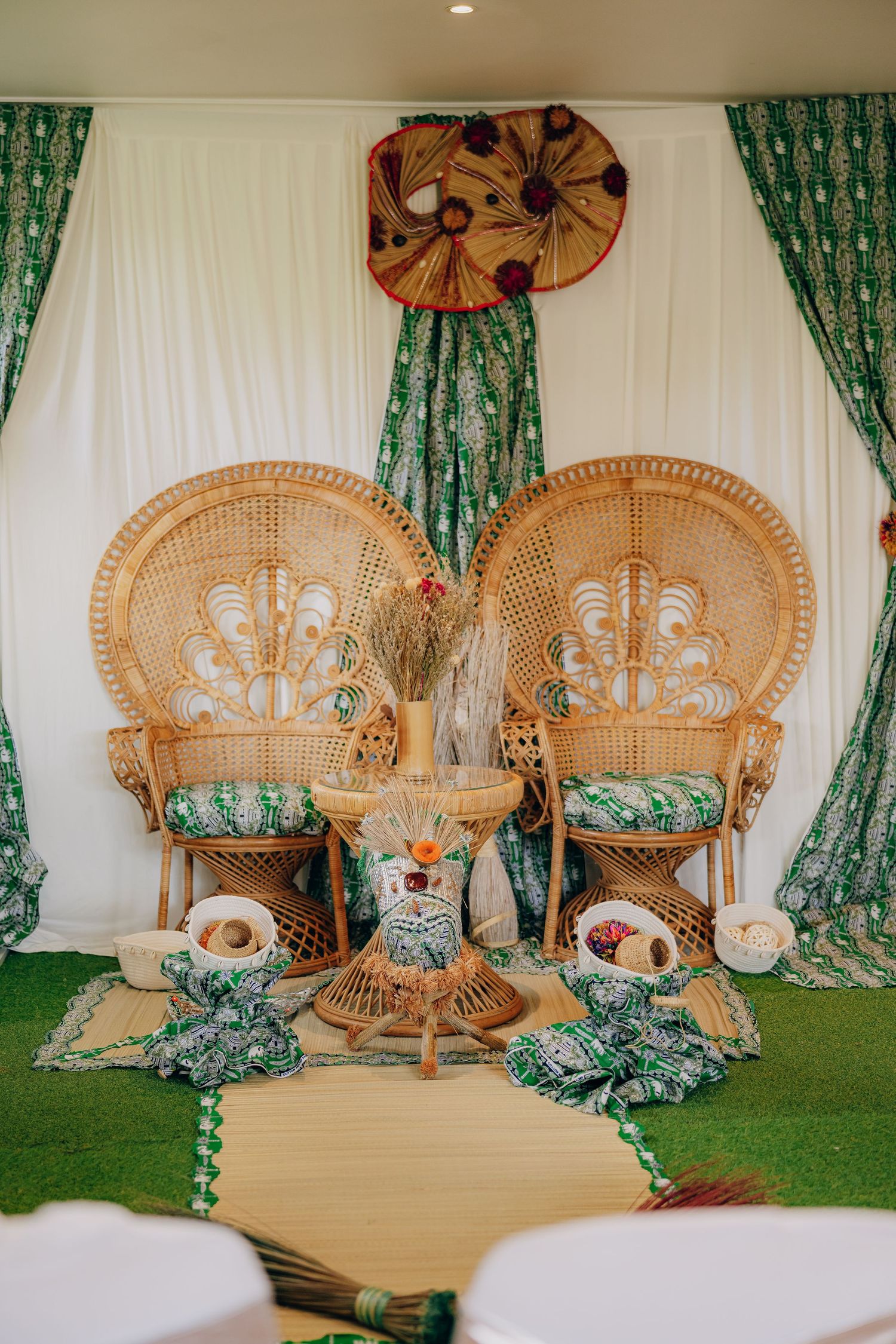 Elegant wedding decor setup with peacock chairs, green curtains, and floral arrangements against a white backdrop.