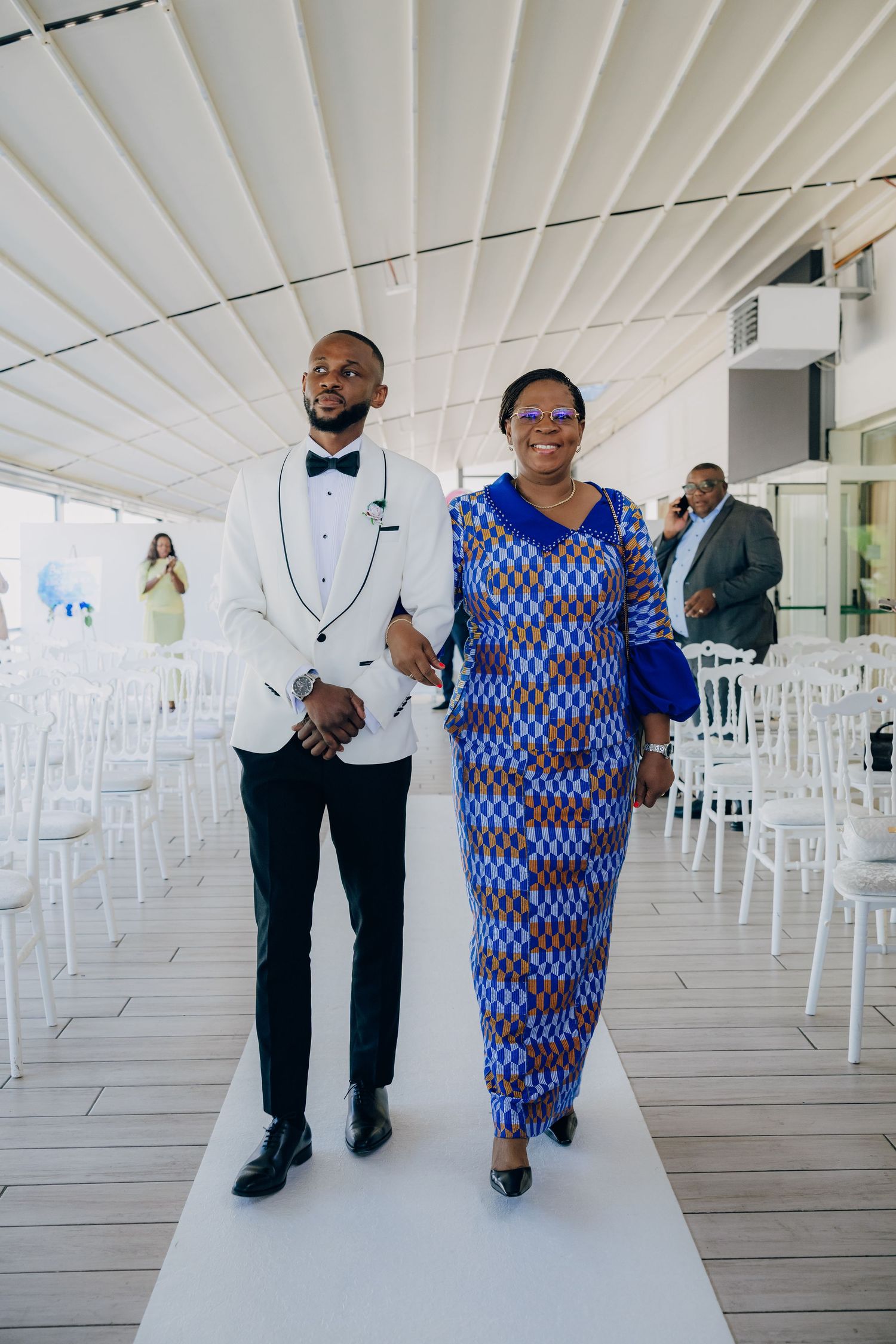 A couple walks down a white aisle, with the woman wearing a vibrant blue patterned dress and the man in a white tuxedo jacket.