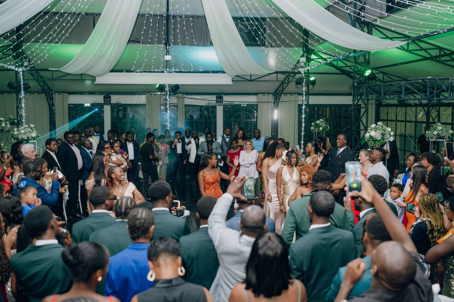 Panoramic view of a wedding reception with guests dancing under green lighting effects.