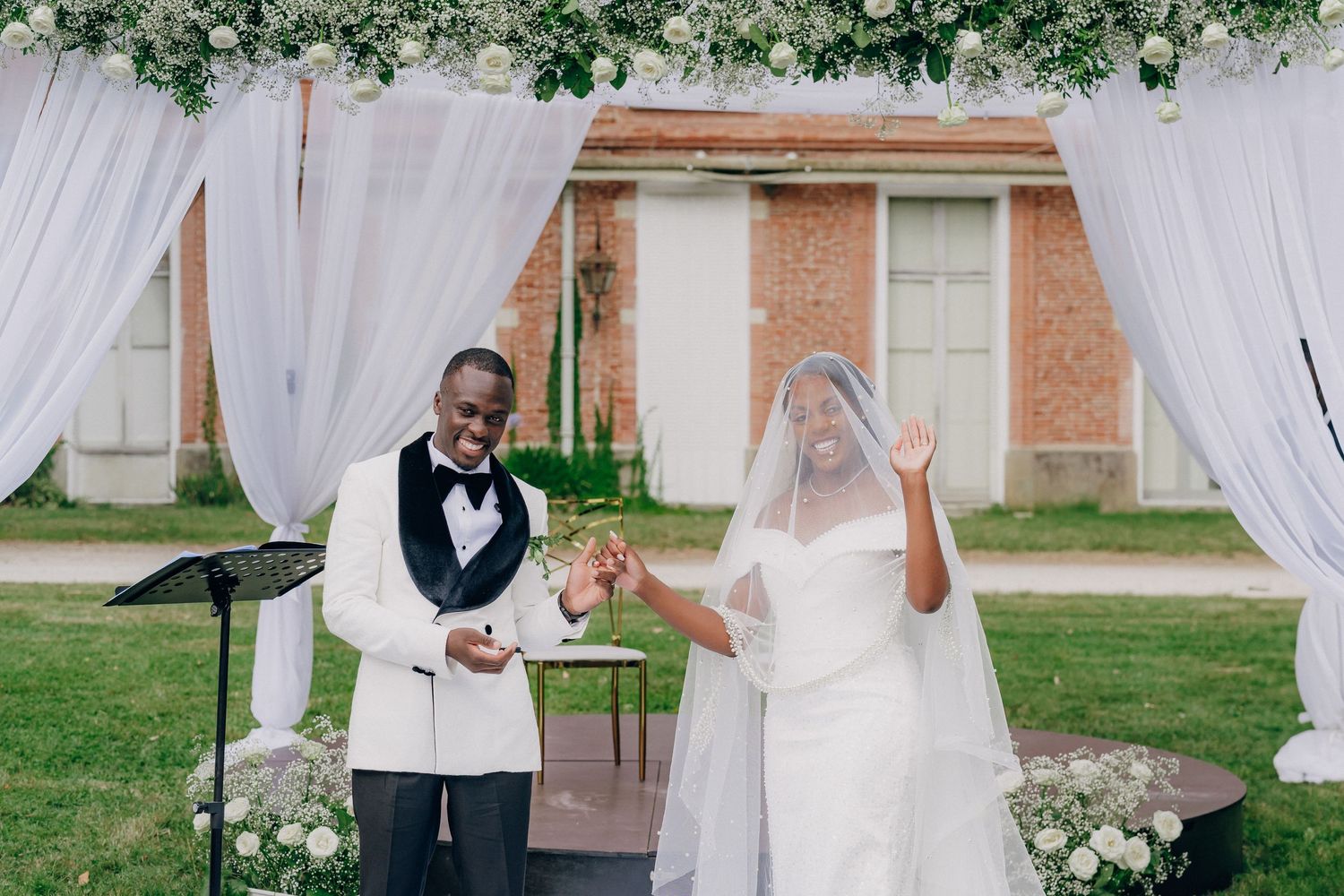 Outdoor wedding ceremony under floral arch with white draping and guests watching the proceedings.