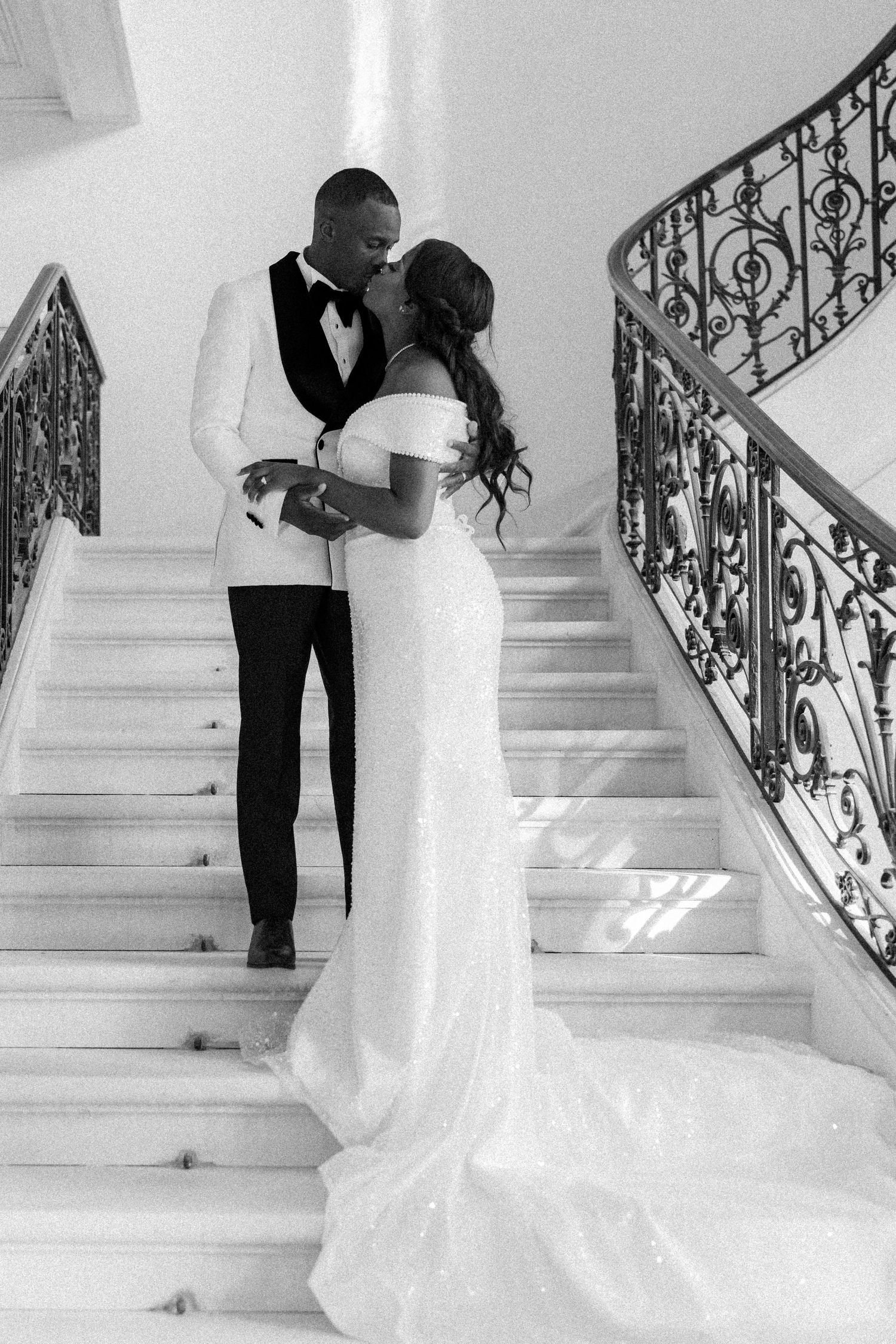 A black and white wedding portrait of a couple sharing an intimate moment on an elegant staircase with ornate railings.